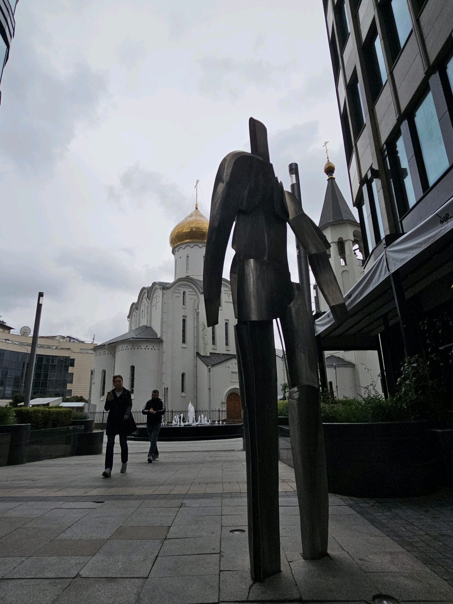 The image depicts an outdoor urban scene under an overcast sky, featuring a modern building with a glass facade and a traditional church with a golden dome in the background. In the foreground, a large, abstract metal sculpture stands prominently. Two individuals are walking on a paved pedestrian walkway. The church appears to be the Church of the Resurrection of Christ (also known as the Church of the Holy Resurrection) located in Moscow, Russia, which is situated next to the modern Office Center "Dominion Tower". The architecture suggests a juxtaposition of old and new urban elements. The lighting is diffused, consistent with cloudy weather.