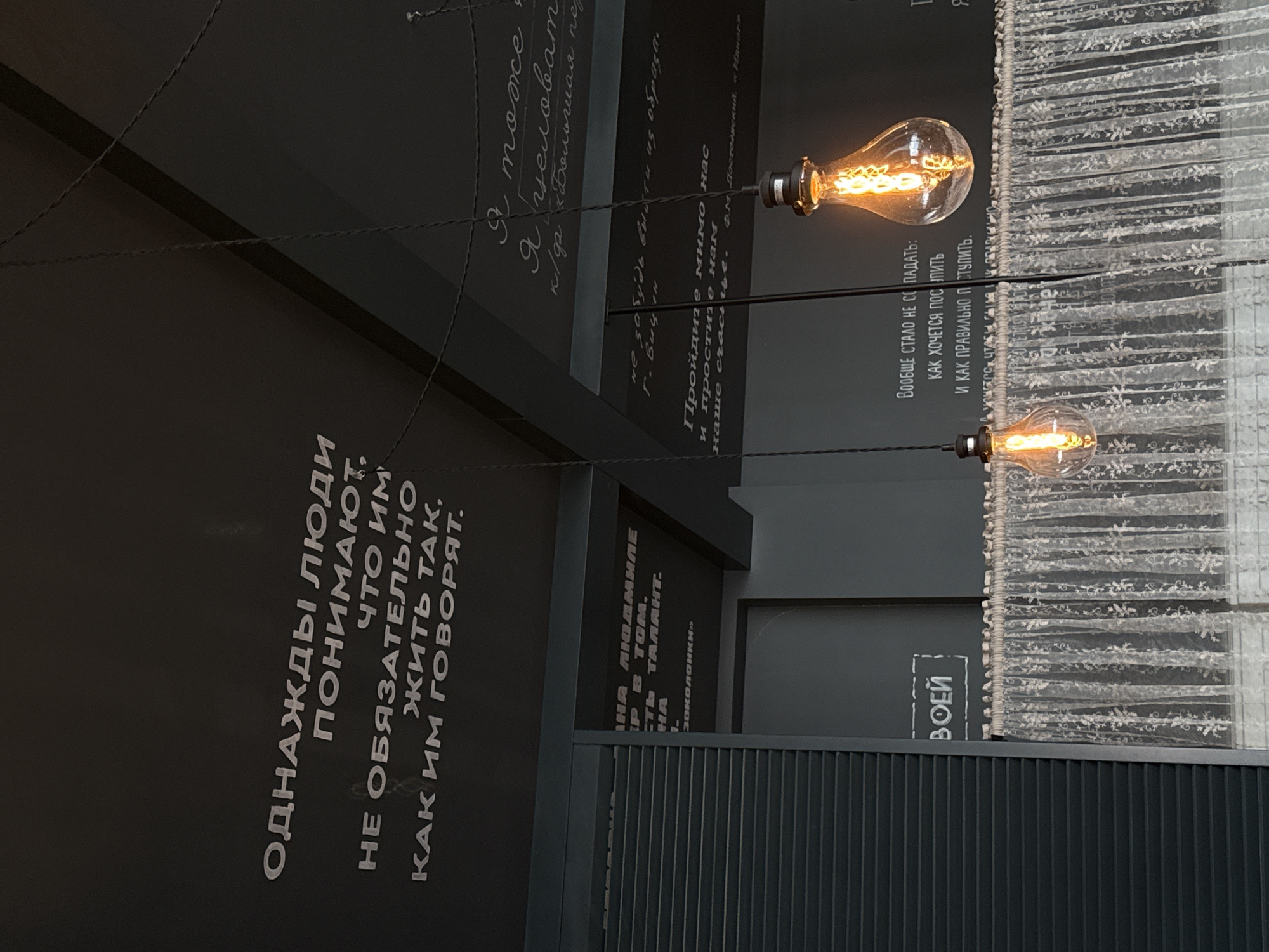 This indoor shot features hanging lightbulbs, dark walls with white Russian text, and a decorative curtain.
The location is suggested by the text on the wall, which includes a reference to Fyodor Dostoevsky and the phrase "Воей," which may refer to a place or person. Given the language and a mention of Dostoevsky, a Russian setting is likely.