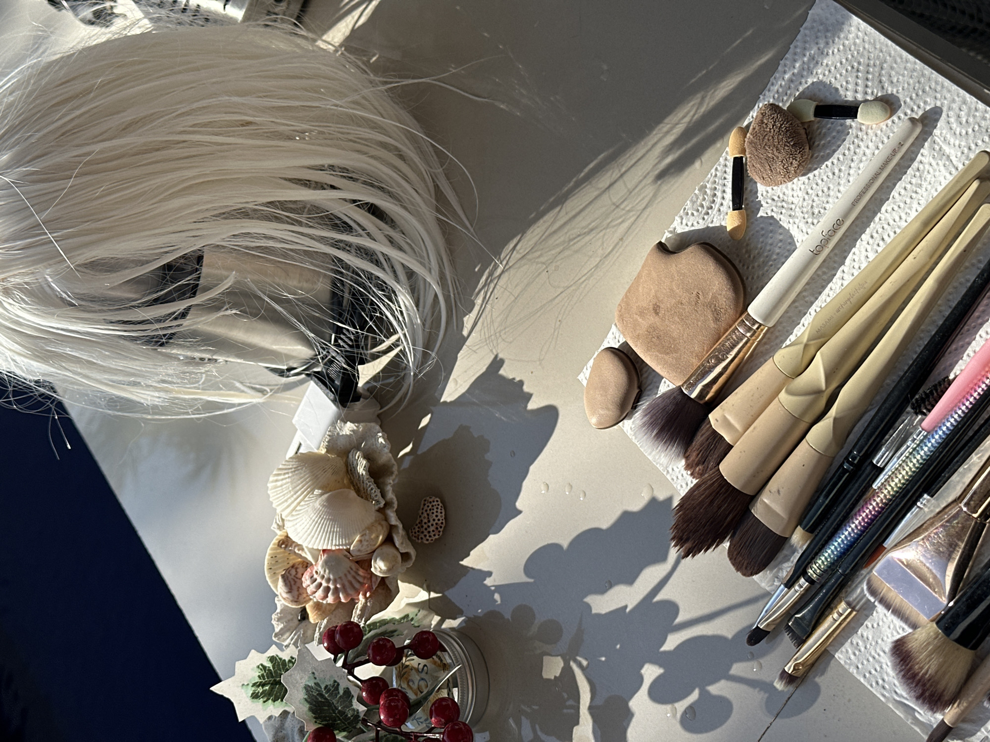 A flat lay composition features a collection of makeup brushes and sponges arranged on a white paper towel. The brushes vary in size and bristle type, with some exhibiting dark brown and lighter blonde bristles, while others have multicolored, metallic handles. Among the brushes are several small makeup sponges, some with pointed ends and others with rounded, puff-like applicators. A pale beige, heart-shaped makeup sponge lies near the top of the arrangement.
In the background, a white wig with long, flowing hair is partially visible, resting on what appears to be a mannequin head. To the left, a decorative sprig with artificial red berries and green leaves sits in a small metallic container. A cluster of seashells, including spiral and fan shapes, is arranged behind the berry sprig. The scene is illuminated by strong sunlight, casting distinct shadows across the surface, which appears to be a light-colored, possibly outdoor table. Several water droplets are visible on the surface.
The image was captured in North Kuta, Indonesia, suggesting an outdoor setting, likely a patio or poolside area given the shadows and the overall bright, natural lighting. No people are present in the image.
