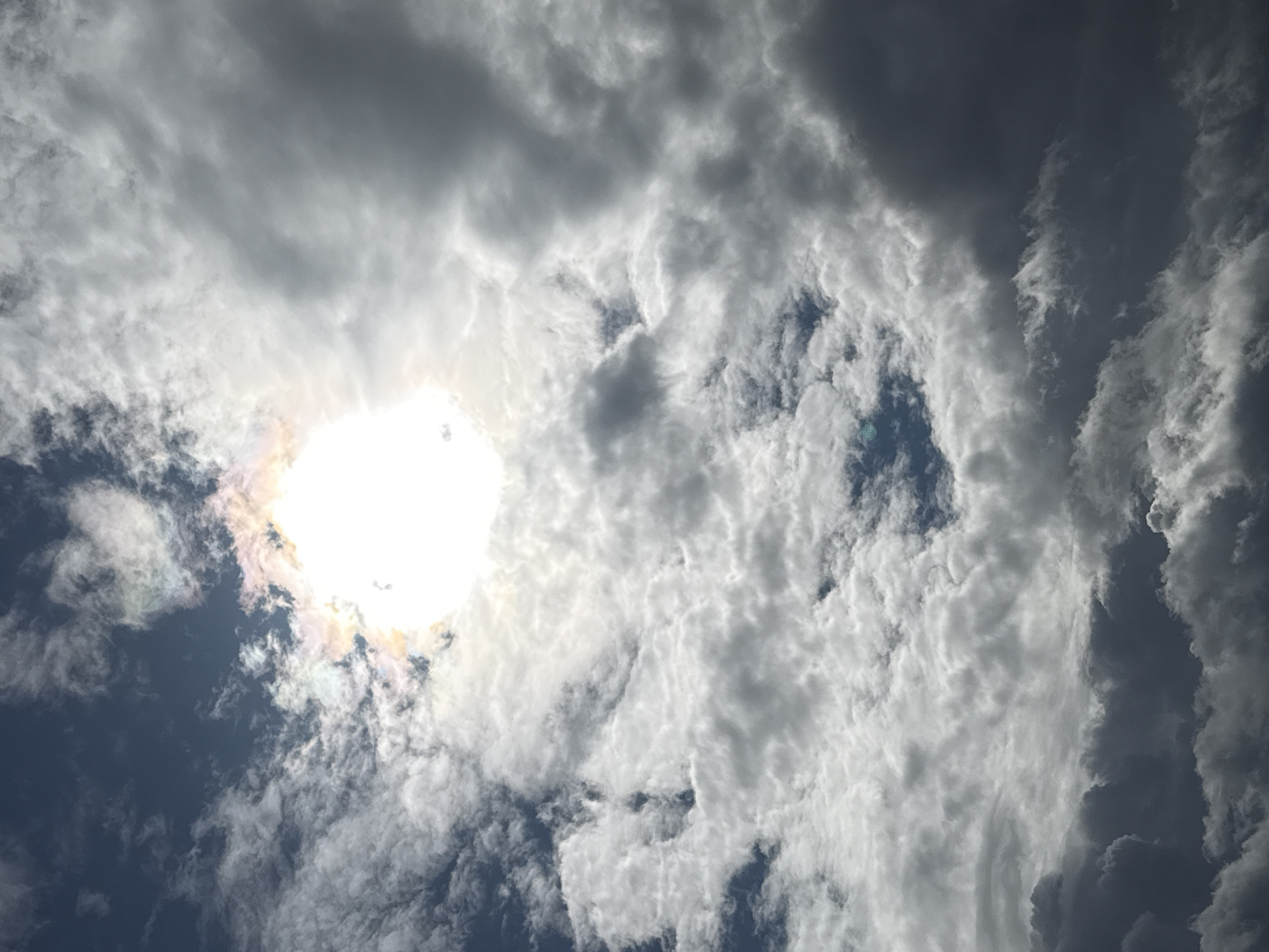 The image shows a sky filled with a mixture of white and dark gray clouds. The sun is visible in the upper center of the frame, partially obscured by bright white clouds. A faint rainbow-like halo is visible around the sun. The sky is a deep blue in some areas, peeking through the clouds. The overall impression is of a dynamic and dramatic sky, possibly before or after a storm. There are no people, structures, or any other discernible objects or subjects in the image. The location is stated to be in the city of Galinė, Lithuania, however, there are no visual cues in the image to confirm this or to provide any specific details about the city itself.