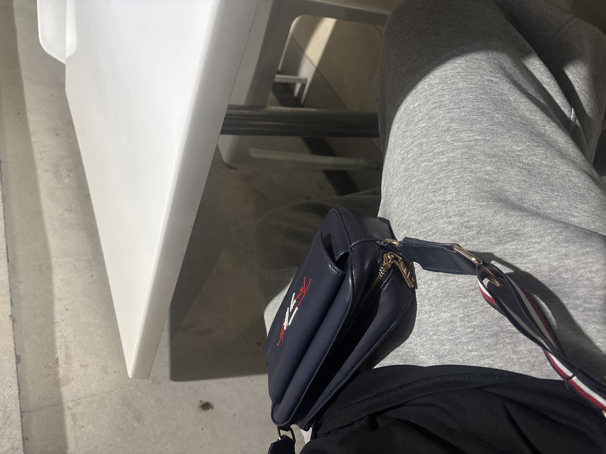 The image presents a close-up, high-angle view focused on a person's lap and surrounding environment. The primary subject is an individual partially visible, wearing light grey sweatpants or track pants. A dark blue shoulder or crossbody bag rests on their lap. The bag has a gold-toned zipper and a distinctive strap featuring red, white, and blue stripes. An embroidered logo on the front of the bag appears to be "TOMMY" or a similar brand identifier. Below the bag, a black garment is visible.
To the left of the frame, a prominent white surface, likely a table leg or part of a chair, is visible. The background consists of a concrete or tiled floor with discernible lines and textures. Shadows are cast across the floor and the grey pants, suggesting a clear light source, possibly natural light, indicating it is daytime. The setting appears to be an indoor or covered outdoor area, potentially a cafe, waiting room, or a public seating space. No specific activity beyond sitting or resting is evident. No direct visual cues within the image indicate the provided context of Yerevan, Armenia, nor are any emotions discernible from the partial view of the person.