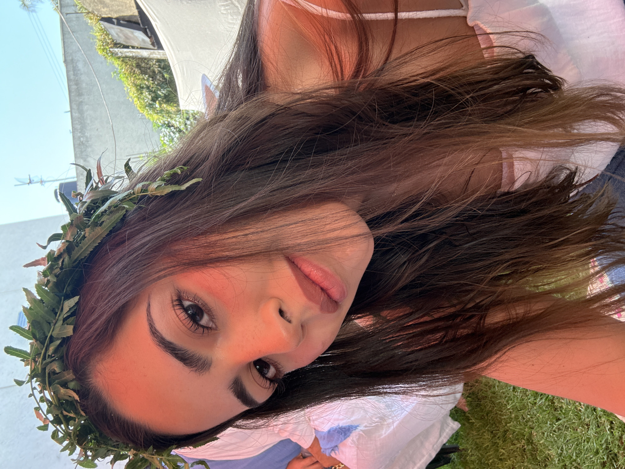 A young woman with long brown hair, adorned with a green leaf crown, poses for a selfie. She has fair skin, dark eyebrows, and brown eyes. She wears a white top with thin straps. The background features a partially visible white canopy, greenery, and a glimpse of a building, suggesting an outdoor setting. The location, Puebla, Mexico, is not discernible from the image.