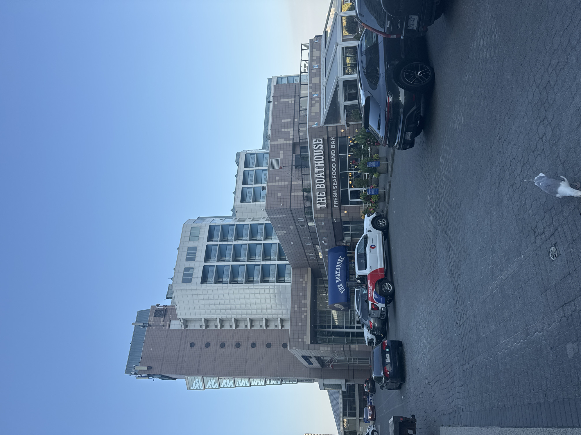 In New Westminster, Canada, an exterior view of The Boathouse restaurant, a seafood establishment, is captured on a clear day. The restaurant is situated at the base of a multi-story building with a modern architectural design, featuring a combination of beige, brown, and white facades with balconies visible on upper levels. A blue awning with "THE BOATHOUSE" prominently displayed in white lettering shades the entrance, below which is signage reading "FRESH SEAFOOD AND BAR". In front of the restaurant, several vehicles are parked in a cobblestone-paved area. These include a white pickup truck with red and blue branding and text reading "WE BUILD BETTER TOGETHER", a dark sedan, a dark SUV, and other vehicles partially visible. A seagull is foraging on the ground in the foreground. The environment suggests an urban waterfront setting, with the clear blue sky indicating fair weather.