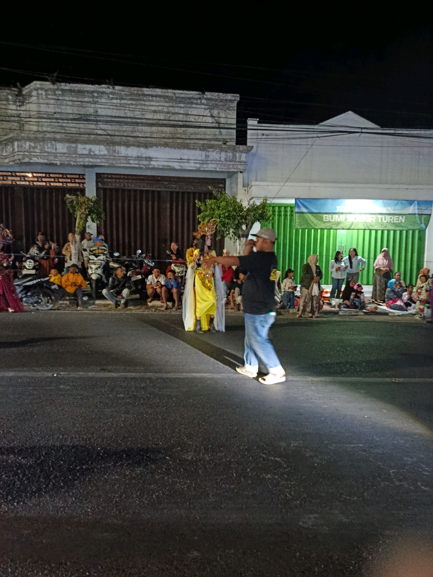 The image depicts a nighttime street scene in Turen, Indonesia, likely during a cultural event or parade. A man in a black t-shirt, blue jeans, and a grey baseball cap is the central subject, captured in mid-stride as he moves across the asphalt road. He appears to be interacting with or gesturing towards a person in a bright yellow, traditional costume with a golden headdress, who is standing on the side of the road. Several other individuals, some dressed in traditional attire, are seated or standing along the roadside, observing the event. Motorcycles are parked behind them, suggesting a public gathering. In the background, a building with a green facade and a banner displaying text that includes "BUMI SUBUR TUREN" is visible. The lighting indicates nighttime conditions, with artificial lights illuminating the scene.