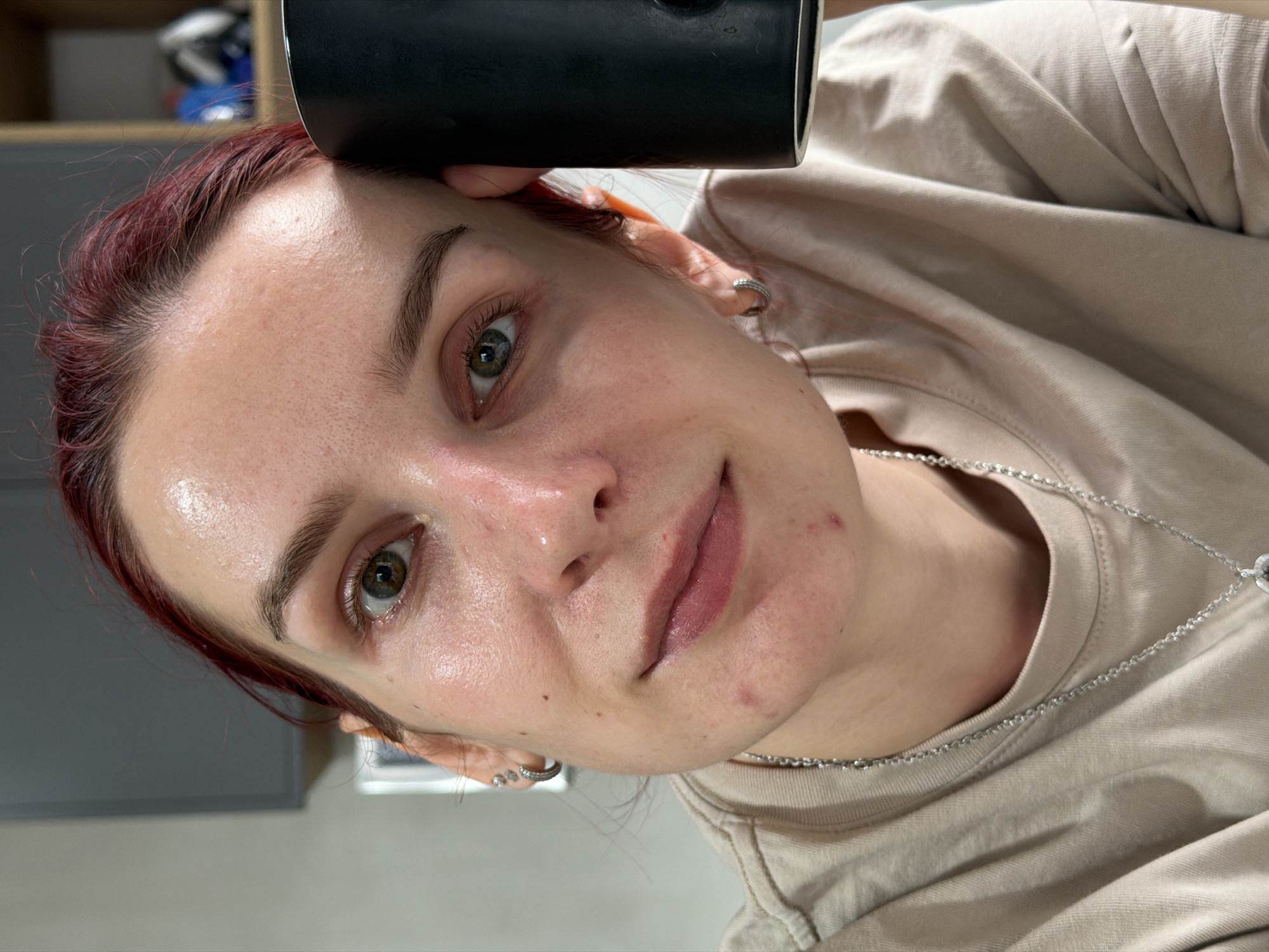 This close-up, tilted selfie captures a person, likely female, in an indoor setting in Chaikovsky, Russia. The subject has reddish-brown hair, fair skin with visible pores and minor blemishes on the forehead and chin, and green/hazel eyes. They are looking directly at the camera with a subtle, gentle smile.
The individual is wearing a light beige short-sleeved t-shirt and a thin silver chain necklace. A small, silver hoop earring is visible in their right ear. A dark, cylindrical object, possibly a cup or part of the camera equipment, is held by the person and partially obscures the top of the frame above their forehead.
The background is softly blurred, showing muted grey tones, indicative of an indoor environment, possibly a home, with hints of cabinetry or shelving in the upper left. The lighting is even, suggesting artificial light or indirect natural light. No text is visible in the image. The overall impression is candid and natural.