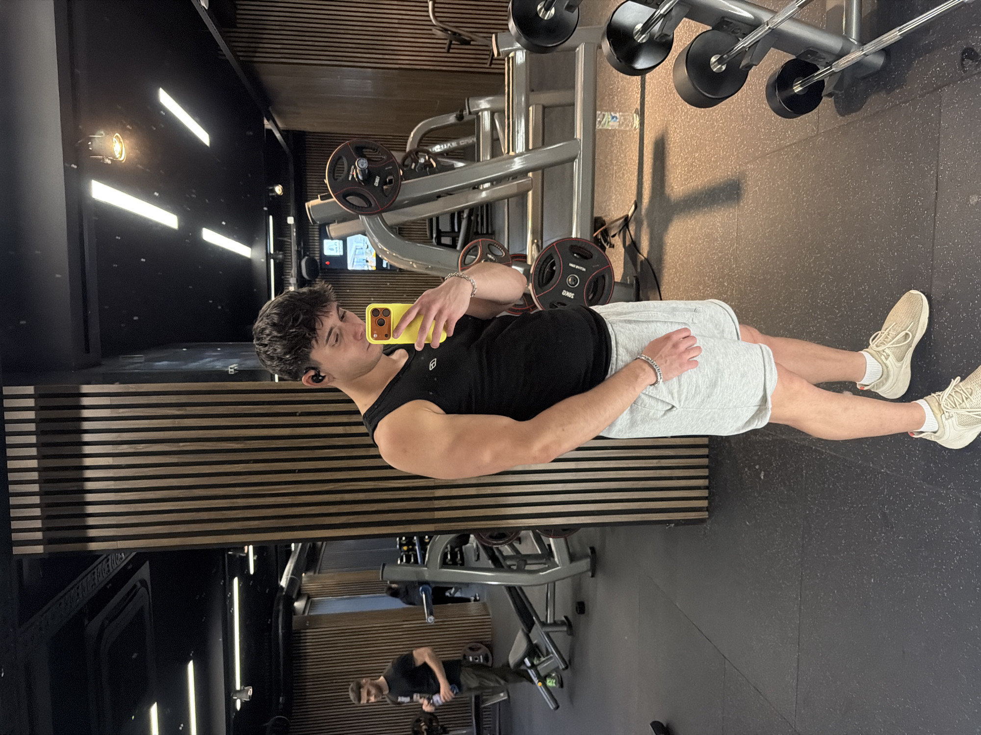 A young man is taking a selfie in a gym. He is wearing a black tank top, gray shorts, and white sneakers. He is holding a yellow smartphone in his left hand. He has dark hair and is wearing an earbud in his left ear. The gym has black walls with horizontal wooden panels, gray equipment, and black rubber flooring. There are weights on the equipment. Another man is working out in the background. The location is likely in a modern, urban gym in Sofia, Bulgaria, given the style of the interior design.
