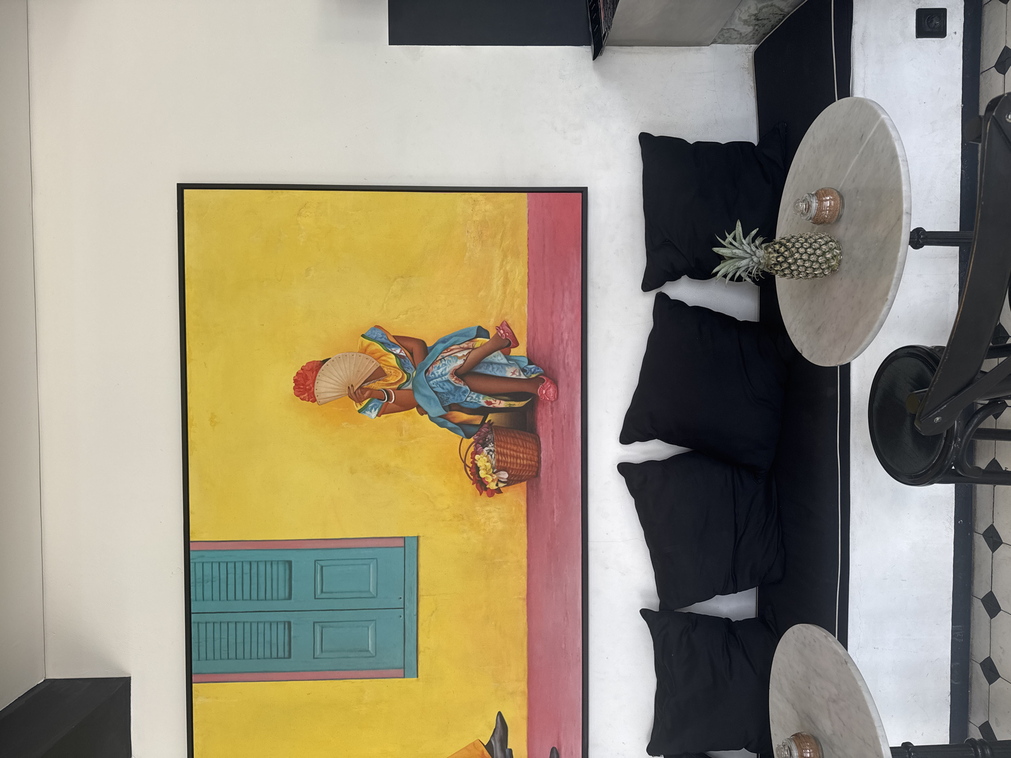 A depiction of an interior in North Kuta, Indonesia. The primary focus is a painting with a black frame depicting a woman holding a fan and basket. The painting features bright yellow and pink sections, and a blue window. Next to the painting is a seating area with black cushions. A marble table stands beside the cushions, adorned with a pineapple and a small jar. Adjacent to the table is a black chair. The floor is white with a black geometric pattern.