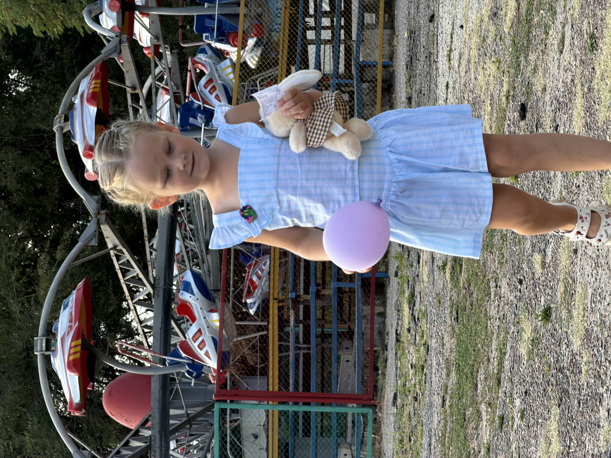A young girl with blonde braided hair, wearing a light blue striped dress, walks towards the viewer outdoors. She is holding a lilac balloon in her left hand and a stuffed rabbit in her right hand, which also holds a receipt. The girl's feet are adorned with white sandals. In the background, a section of a colorful amusement park ride with red and white airplane-shaped seats is visible. The ground is covered with gravel and sparse green vegetation. The location is identified as Kamyshin, Russia, based on contextual information.