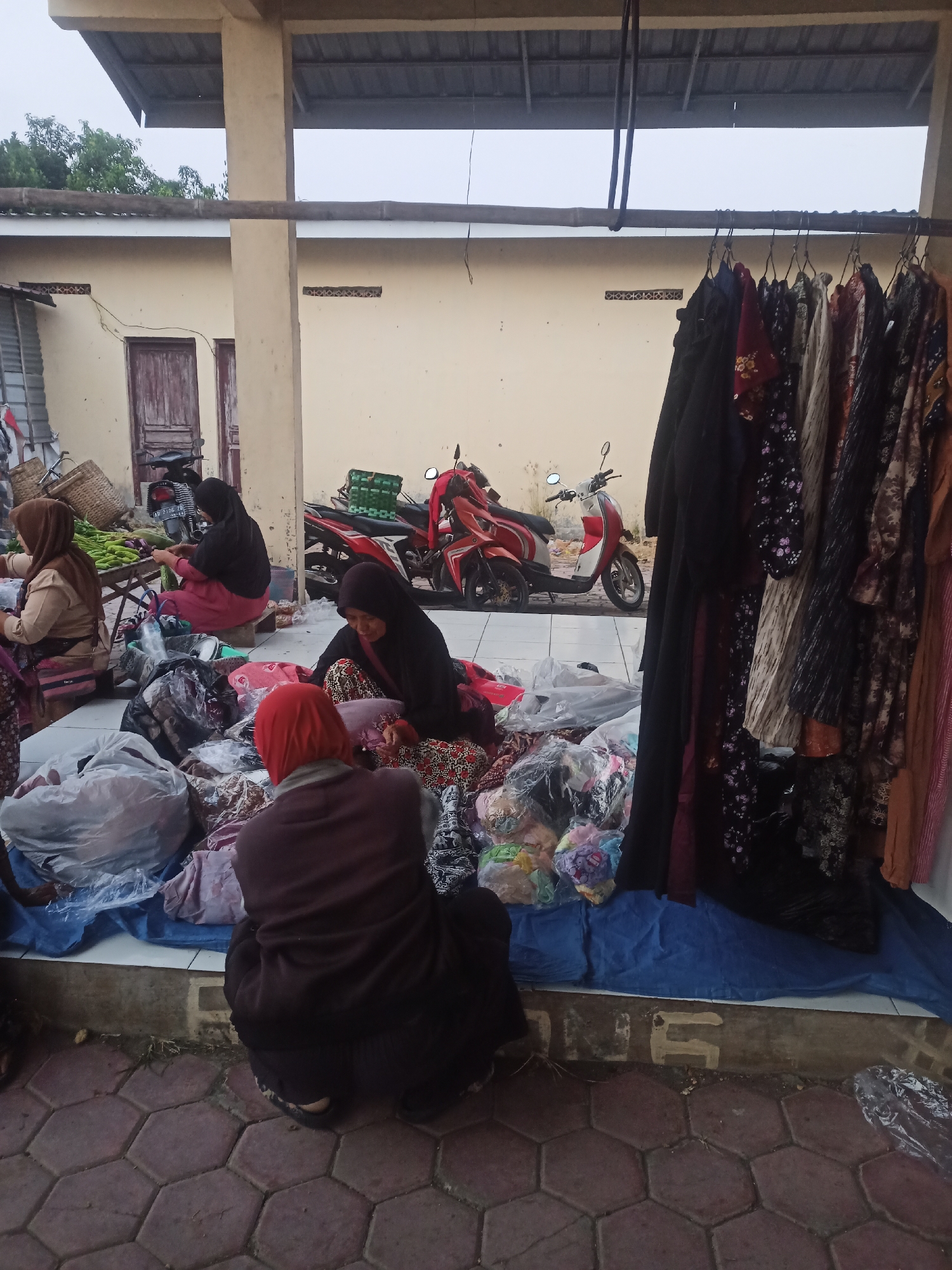 A group of women are gathered around a market stall in Nganti, Indonesia, selling clothing and produce. The women, all wearing hijabs, are interacting with the goods and each other. Some are seated on the ground, while others are on stools. The stall is set up under a covered walkway, with clothing racks displaying various garments and piles of produce on a table. Two red and white motorcycles are parked behind the stall, indicating a common mode of transportation in the area. The ground is paved with hexagonal tiles, and the background shows a light-colored building.