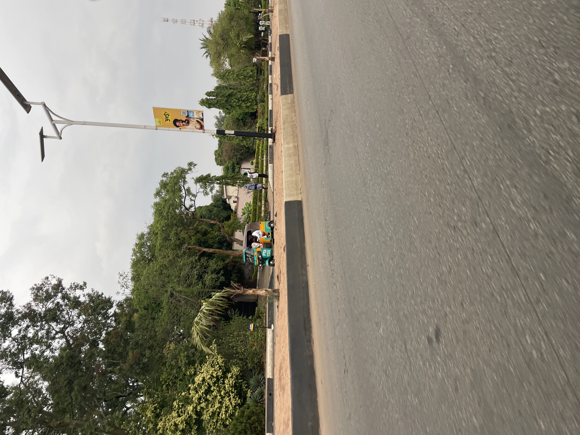 The image shows a street scene in Jos, Nigeria. The foreground is dominated by a grey asphalt road. To the left of the road is a curb and a sidewalk lined with trees and vegetation. A three-wheeled vehicle (a keke) is parked on the sidewalk, near the curb. A light pole with a billboard advertising a product is visible on the left. Further back, a telecommunications tower can be seen. Several people are walking in the distance on the left side of the road. The sky is overcast.