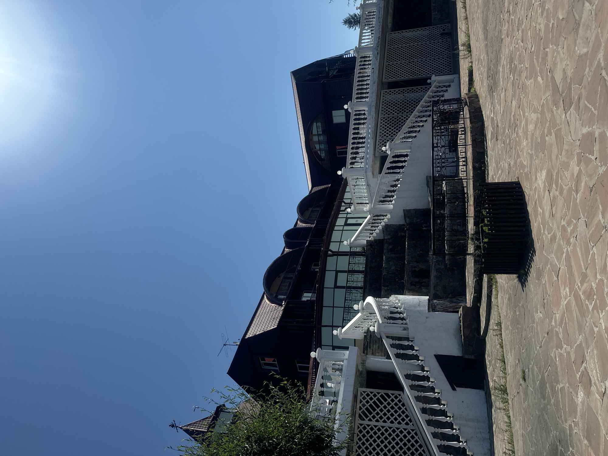 An exterior, low-angle, full shot shows a large, multi-story building with a dark brown, sloped roof against a clear, bright blue sky with the sun glinting in the upper right corner. The building appears to be a hotel or a large restaurant, with several arched windows and balconies visible on the upper levels.
In the foreground, a wide, white staircase with ornate railings ascends towards the building. To the left of the staircase, a portion of a white trellis wall is visible. The ground in front of the staircase is paved with irregular, reddish-brown flagstones, with some patches of grass growing between them. A dark metal fence encloses a small area to the right of the staircase, possibly a planter or a well.
On the far left of the image, a tall, slender tower with a pointed roof and a weather vane peeks out from behind a leafy green tree. The building also has a modern section with large, floor-to-ceiling windows.
This building is the "Listvyanka" restaurant, located in the village of Listvyanka, Russia, on the shore of Lake Baikal. The photograph captures the building on a sunny day, highlighting its architectural features and the surrounding landscape.