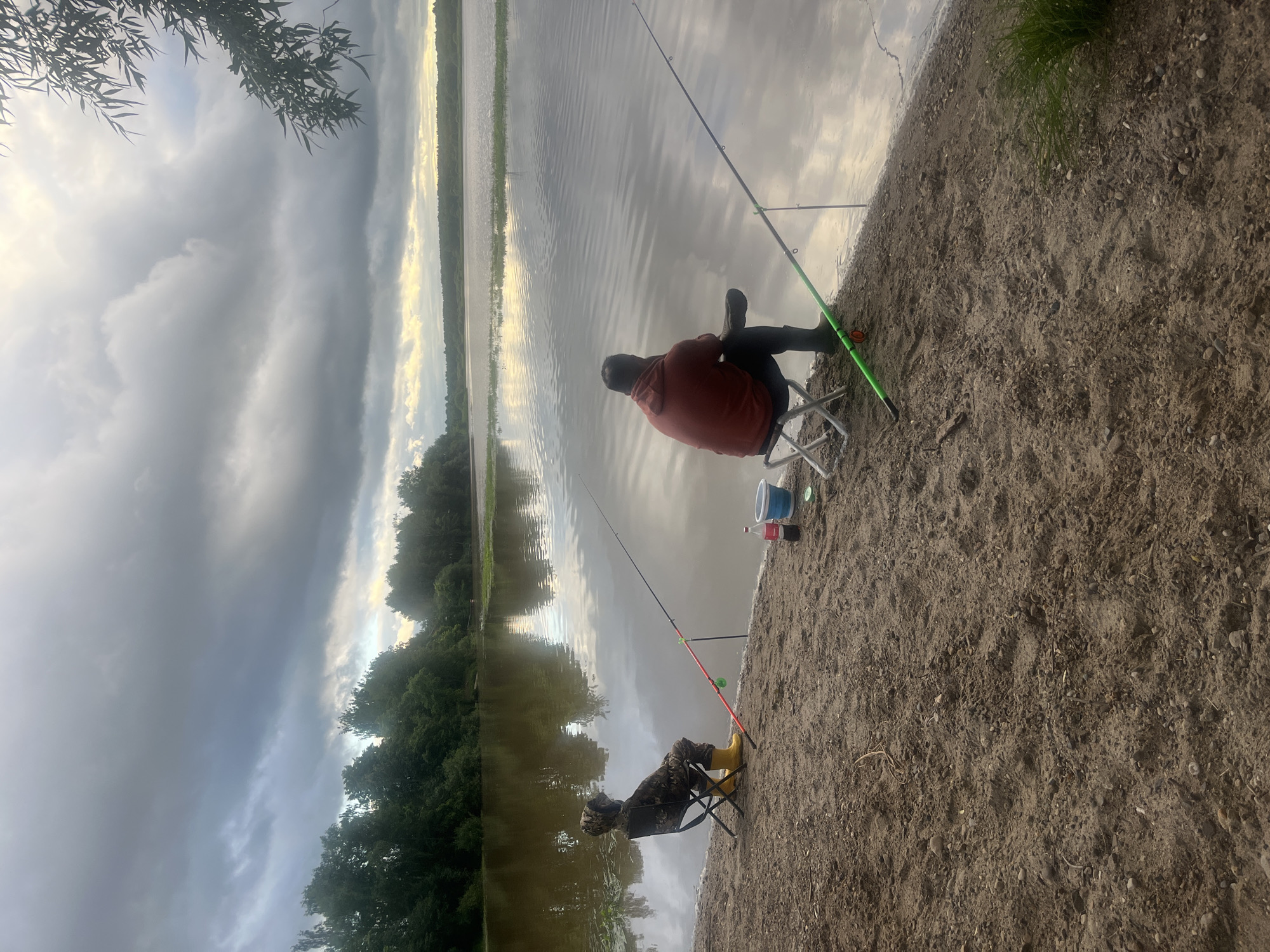 The image depicts two individuals fishing by a calm body of water. The scene is set on a sandy shore, with lush green trees lining the opposite bank of the water. The sky is overcast with dramatic clouds, suggesting it might be late afternoon or early evening, with a hint of sunlight breaking through on the horizon.
One person, wearing an orange hoodie and dark pants, is seated on a folding chair, holding a fishing rod. Another person, camouflaged in what appears to be fishing attire and yellow boots, is also seated on a folding chair and holding a fishing rod. Their fishing rods are propped up, indicating they are waiting for a bite. Various fishing gear, including a bucket and a bottle of Coca-Cola, are placed near the shore.
The overall atmosphere is one of peaceful recreation. The calm water reflects the sky and trees, creating a serene and natural environment. The location is identified as Klepiki, Russia. There is no visible text in the image.