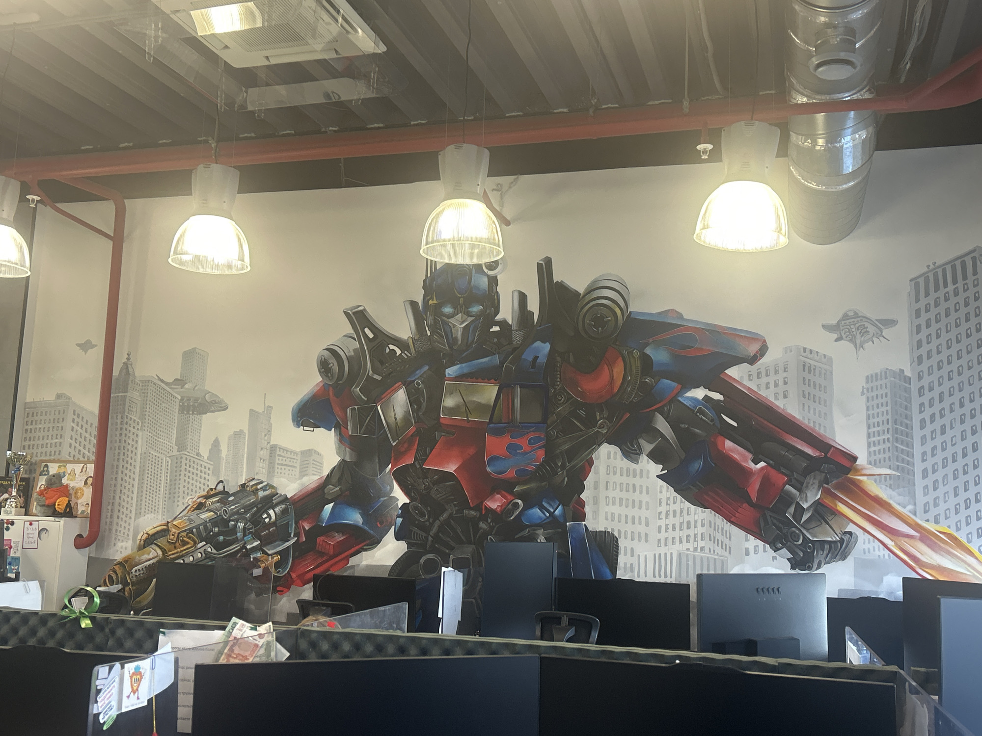 This media file shows a large mural of Optimus Prime, the iconic Transformer character, painted on a wall in what appears to be an office setting. Optimus Prime is depicted in a dynamic pose, with his signature red and blue armor, flames on his chest, and a weapon in his right hand. He is surrounded by a cityscape background, with skyscrapers and flying vehicles, suggesting a scene of action and urban warfare.
The mural is illuminated by several pendant lights hanging from the ceiling, which also features exposed ductwork and industrial elements. In the foreground, cubicle dividers and computer monitors are visible, indicating the space is likely a workplace. Some papers and office supplies are scattered on the desks, and a trophy is displayed on a shelf to the left.
The overall impression is one of a modern, vibrant office space that incorporates pop culture elements to create an engaging atmosphere. The mural itself is highly detailed and colorful, bringing the character to life. There is no discernible time of day or weather, as the scene is indoors. However, the choice of a powerful, heroic figure like Optimus Prime could evoke feelings of inspiration or excitement among the office occupants. Some faint Cyrillic text is visible on papers in the foreground, but it is not legible enough to be deciphered. The location context is identified as Perm, Russia.