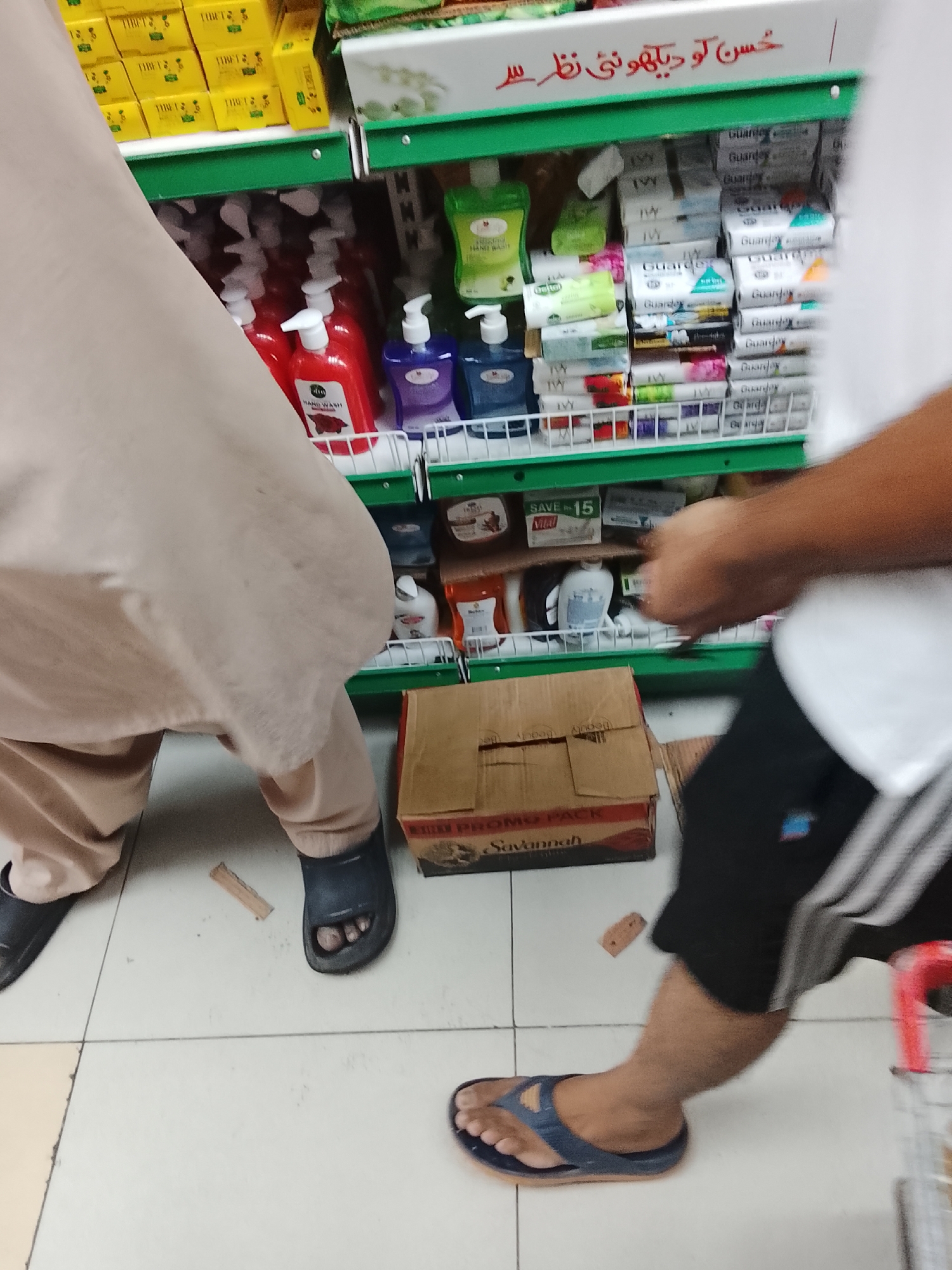 The image appears to be taken inside a retail store in Gulgasht Colony, Pakistan. A low-angle shot shows two people and shopping cart alongside shelves stacked with items such as hand wash, soap, and other household products. The shelves are green and white. One person is wearing a light-colored outfit and dark sandals and bending. The other person is wearing a white shirt and black shorts with white stripes and is wearing blue sandals. A brown cardboard box labeled "Savannah" is on the floor between the two. The floor is tiled. Text in Urdu is visible above the shelves.