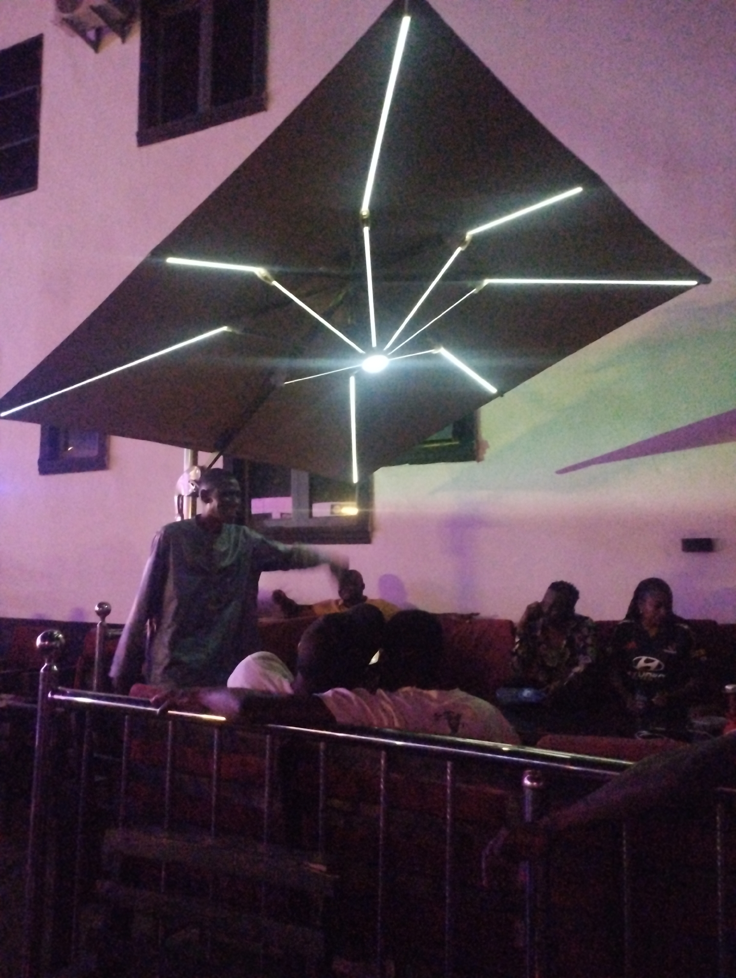 The image shows an indoor lounge scene, potentially in Tumfure, Nigeria. The foreground contains a wrought-iron style railing. Behind it, a group of people are seated on a reddish-brown cushioned bench. One man is standing and gesturing with his right hand. Above, a large, dark, square umbrella with integrated LED lighting is visible. The walls are painted white with visible windows and an air conditioning unit. The lighting in the room is dim, with a purplish hue.