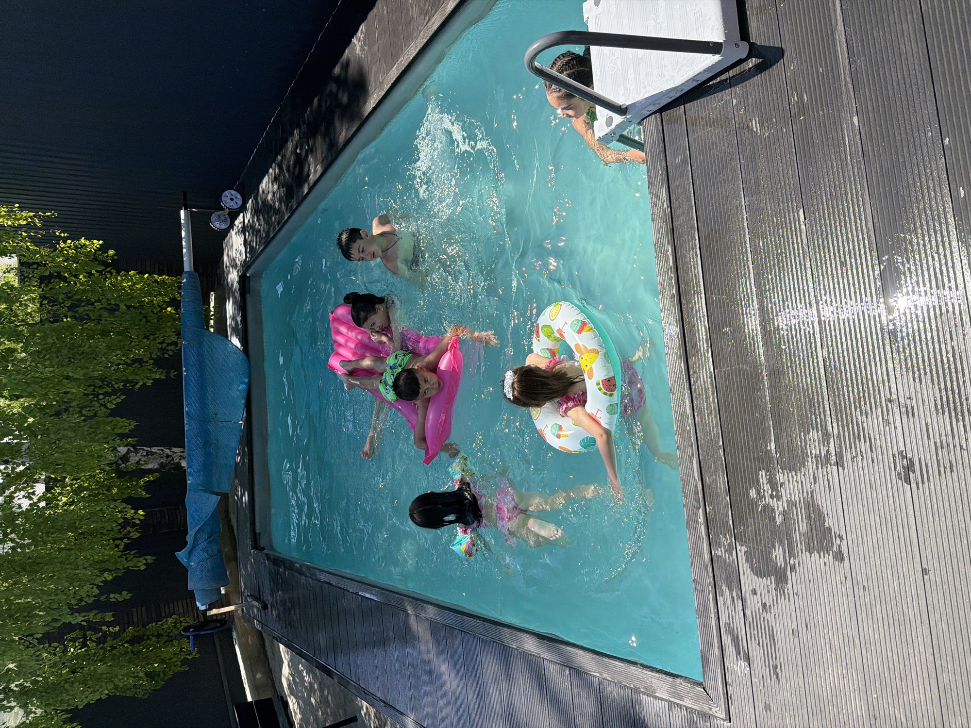 The image depicts a rectangular swimming pool, likely in a backyard, surrounded by wooden decking. Several children are in the pool, some using inflatable floats (one pink, one patterned). One child is partially visible at the pool ladder. The pool is filled with clear blue water. A tree is visible on the left side of the frame. A pool cover roller is located near the tree and the pool. The exact location cannot be determined to be in Goryachevodsky, Russia, based on this image alone. Further contextual information would be needed.