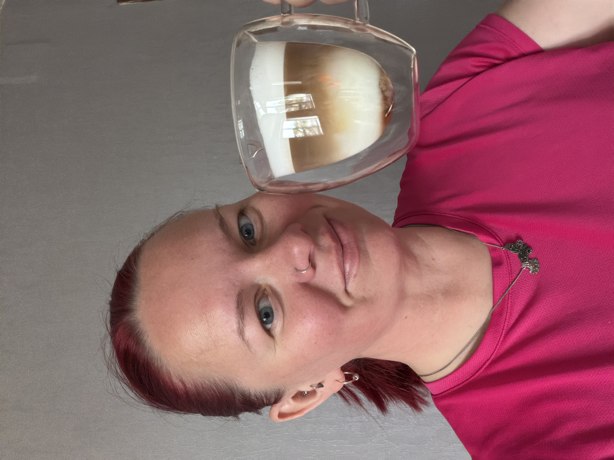A woman with light skin, red hair, blue eyes, and a nose piercing is holding a glass cup containing a layered beige and white beverage. She is wearing a pink shirt and a necklace with floral accents. The background is a plain gray wall. The location, Chaikovsky, Russia, cannot be determined from the provided image.