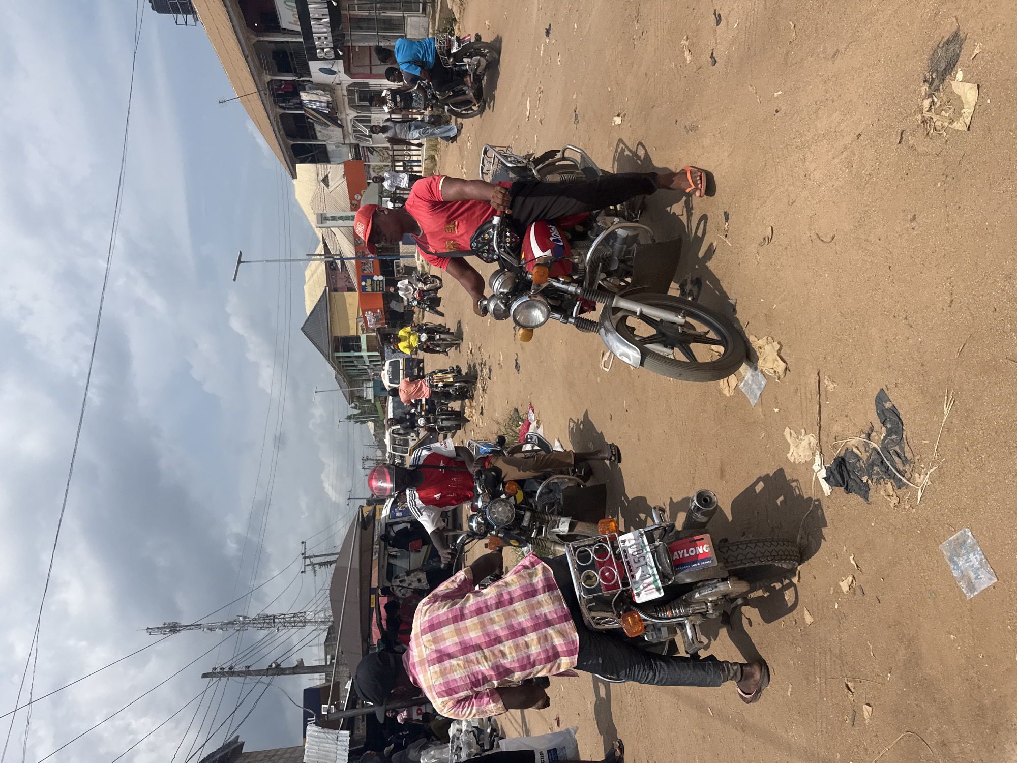 This photograph captures a busy daytime street scene in Ihiagwa, Nigeria, characterized by numerous people and motorcycles. The setting appears to be an unpaved, dusty road lined with commercial buildings and residences.
In the foreground and midground, several men are seated on motorcycles, suggesting they are either waiting, parked, or navigating traffic. A prominent figure on the right is a man wearing a red cap, a red t-shirt, dark trousers, and orange flip-flops, sitting on a red and silver motorcycle. He has a patterned waist bag. Next to him, seen from behind, is another man in a plaid shirt (pink, yellow, and white checks), dark pants, and brown shoes, also on a motorcycle equipped with a rear rack. Further down the street, more individuals are visible on motorcycles, including one man in a bright blue shirt.
The background features a row of modest buildings, some with corrugated roofs and others with flat concrete structures. Utility poles with a dense network of overhead wires run along the street, indicating active infrastructure. A large black water tank is visible on the rooftop of a distant building, and a tall lattice communication tower or similar structure rises against the sky.
The ground is a sandy, light brown dirt road, showing signs of wear and tear, with scattered debris including plastic bags and other refuse. The strong sunlight casts distinct shadows, suggesting a time of day around midday or early afternoon. The sky is partly cloudy, a mix of blue and light grey.
Visible text includes "AYLONG" and "EXPLORE YOUR DESIRE" on a license plate frame on the motorcycle in the foreground. The license plate itself starts with "LAG Q0133," likely indicating Lagos registration. The red motorcycle in the midground has "OYO" faintly visible on its front fender.