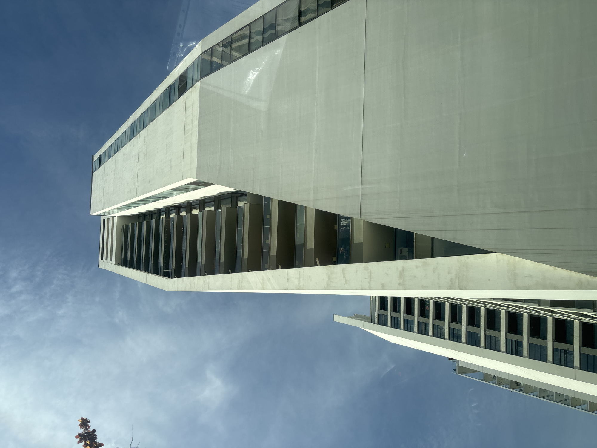 The image shows a modern architectural building against a blue sky. The building is primarily white with sections of glass windows. The structure is angular with distinct geometric shapes. The photograph is taken from a low angle, looking upwards. There are other similar buildings, also modern, in the distance. The location in the city is unknown.