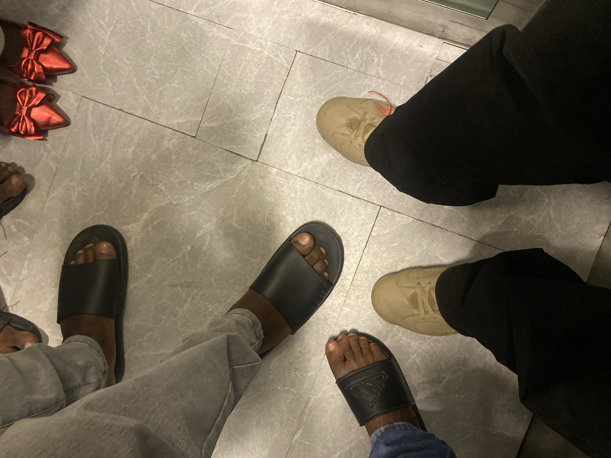 This image shows a top-down view of four people's feet wearing various footwear. The individuals are standing on a grey tiled floor. In the upper left, one person is wearing black slide sandals and light grey pants. To their right, another person is wearing bright red pointed-toe heels with large red bows. In the lower left, a third person is wearing black slide sandals and appears to be wearing blue denim. Directly to the right of them, a fourth person is wearing light brown sneakers and black pants. The setting appears to be indoors, possibly a retail or public space, given the tiled flooring. The specific location in Sabon Gari West, Nigeria is not discernible from the visual information provided in the image.
