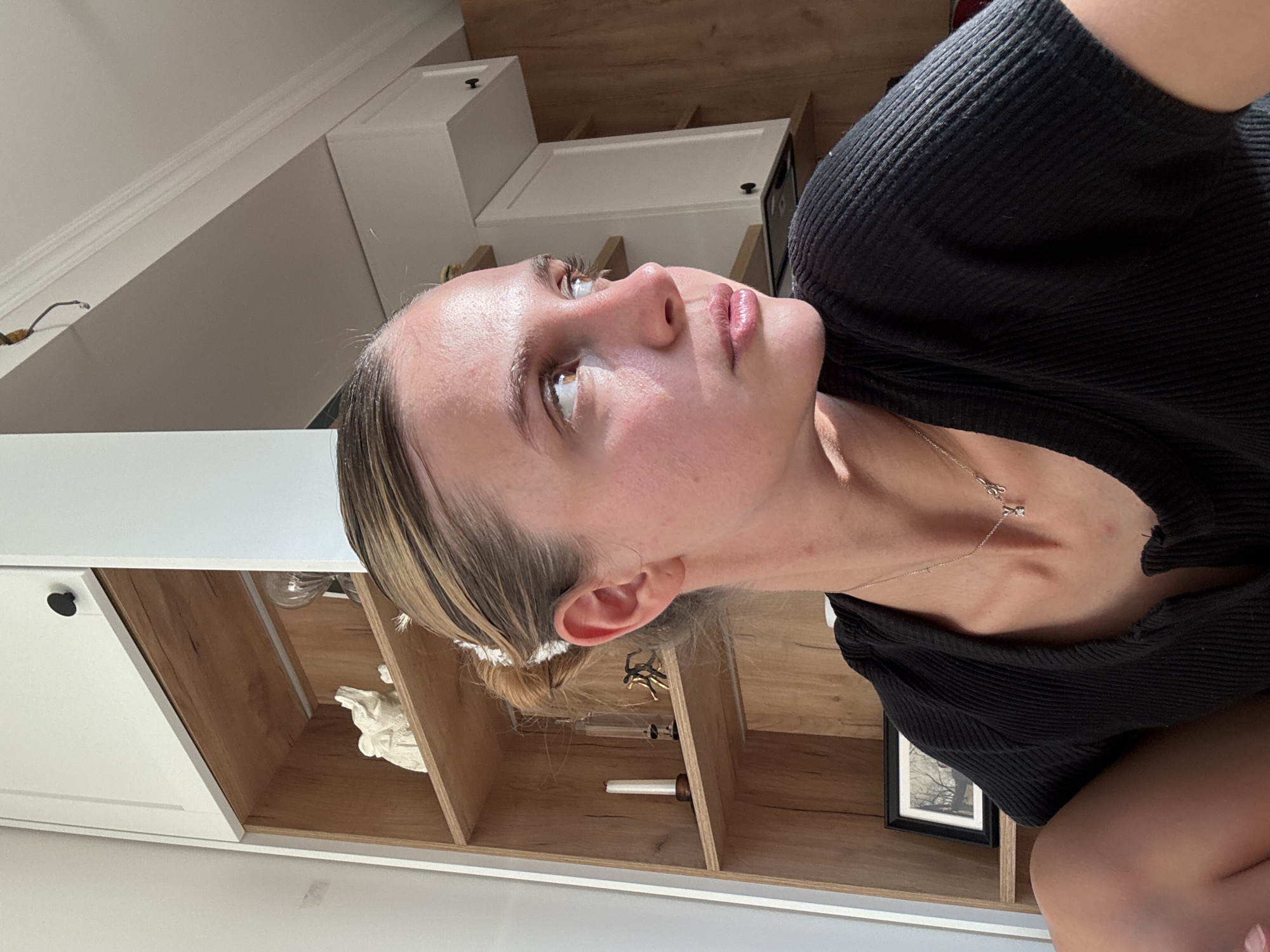 A woman with fair skin, blonde and brown hair pulled back, and wearing a black ribbed top is oriented in a near-vertical position in the frame. She is looking upwards and to the left. A silver necklace with a small pendant is visible around her neck. The background consists of a shelf unit with wooden shelves and white cabinets. Various decorative items are arranged on the shelves. A black-framed photograph is also visible on one of the shelves. The environment appears to be an interior space, possibly a home.
