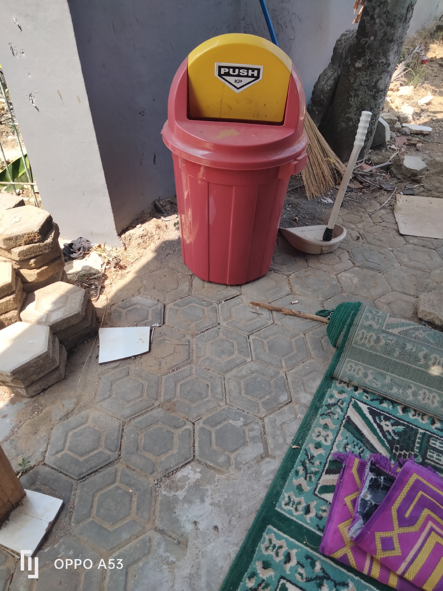 A red and yellow trash can sits on a gray, hexagonal-tiled outdoor area in Badung, Indonesia. A broom and dustpan are leaning against the trash can. Several hexagonal concrete pavers are stacked to the left of the trash can. A green rug with a decorative pattern is partially rolled out on the ground to the right, with two smaller purple mats placed on top of it. The area appears to be a yard or outdoor workspace, with a concrete wall and a tree trunk visible in the background. Sunlight casts a shadow across the scene. The OPPO A53 logo is visible in the bottom left corner.