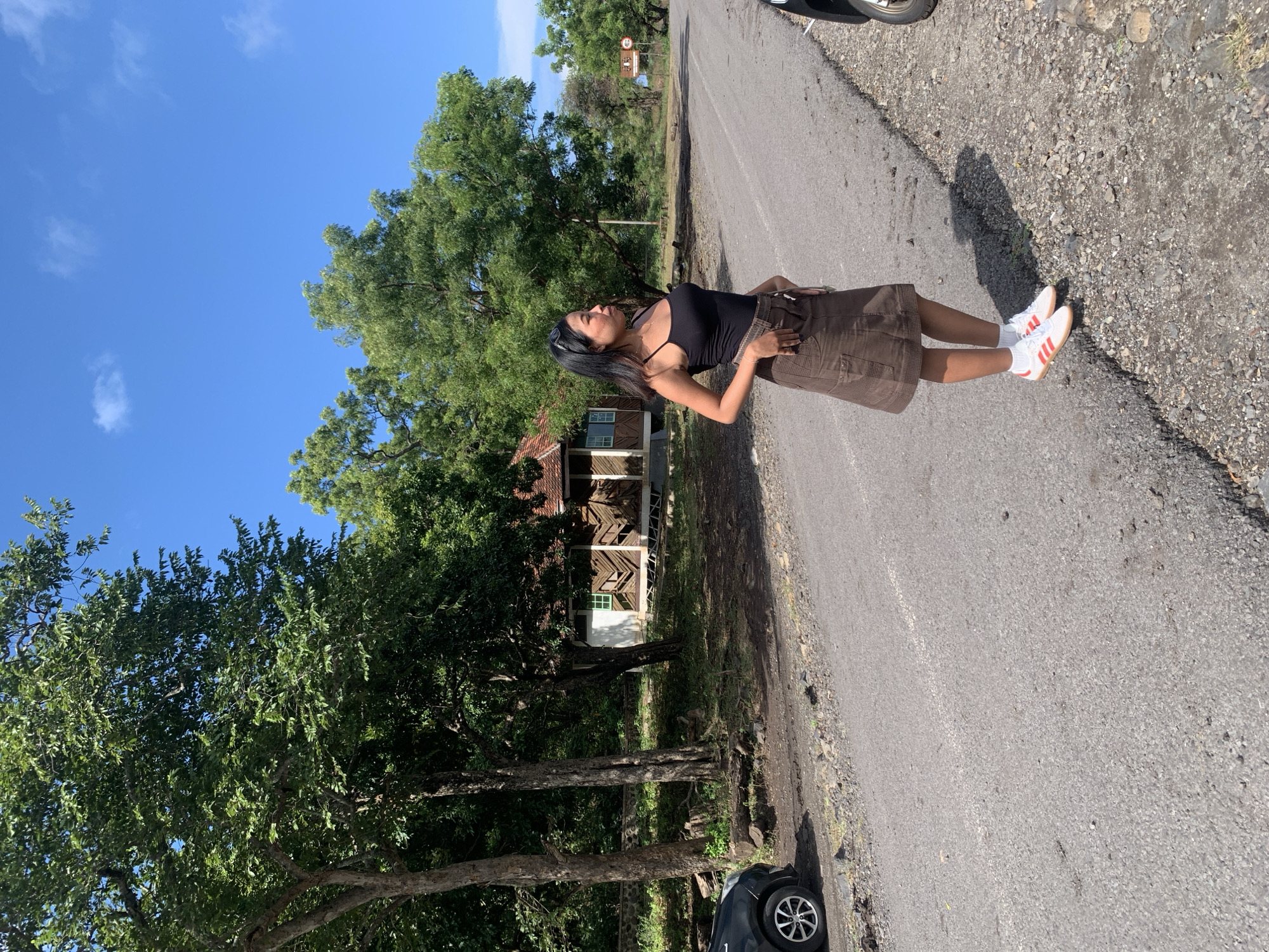 A woman stands on a paved road, gazing upwards. She wears a black tank top, brown skirt, and white sneakers with orange stripes. The road borders a grassy area with trees. A building is partially visible through the trees. The sky is blue with scattered clouds. Part of a vehicle is visible on the side of the road. Location: Unknown, Indonesia.