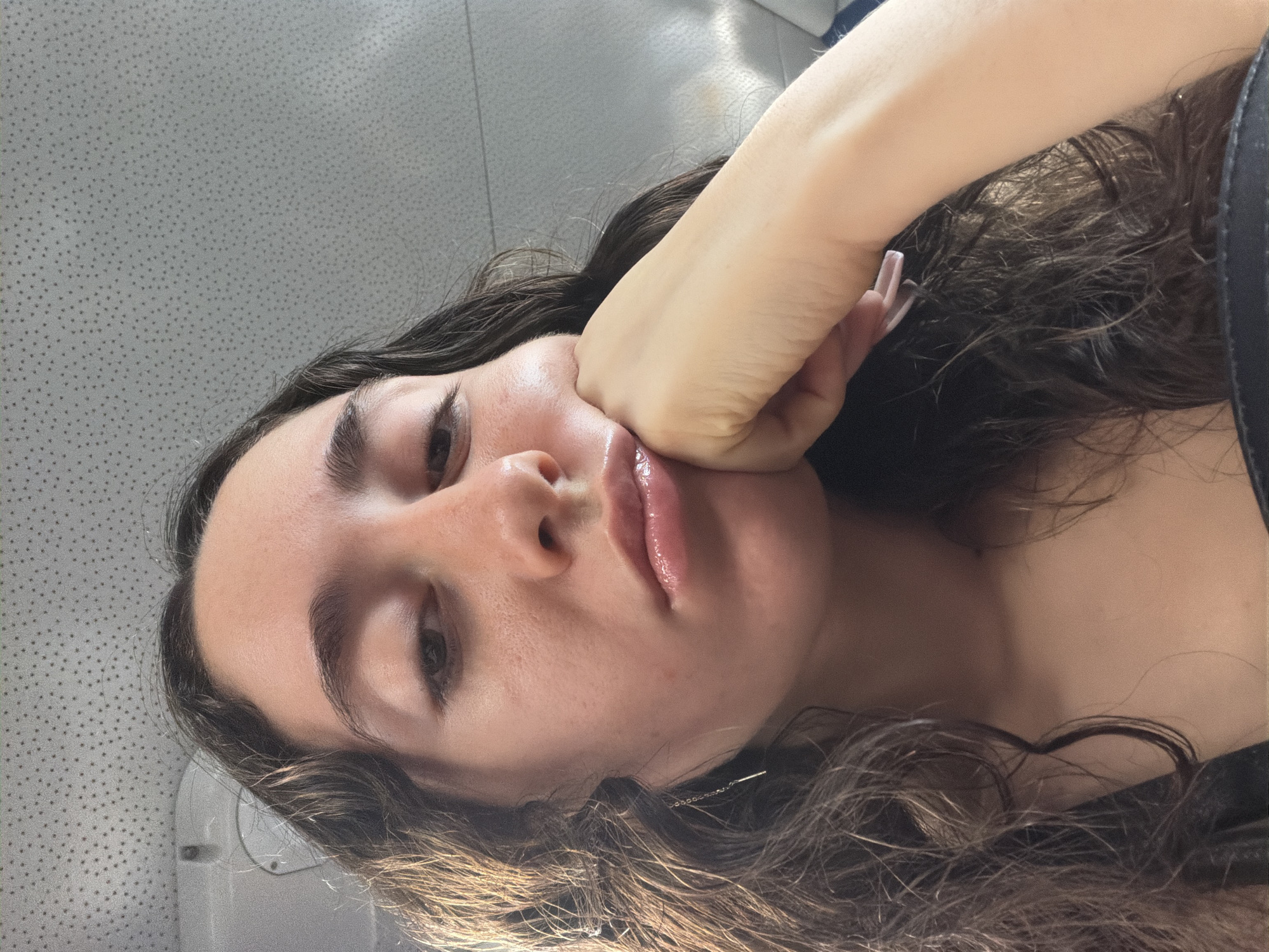 A woman with long, wavy brown hair is photographed from a low angle, facing the camera. Her right hand is pressed against her cheek, causing her face to appear slightly distorted. She has dark eyebrows, brown eyes, and full lips. A small silver earring is visible near her hair. The background is a mottled gray surface. The location is unknown.