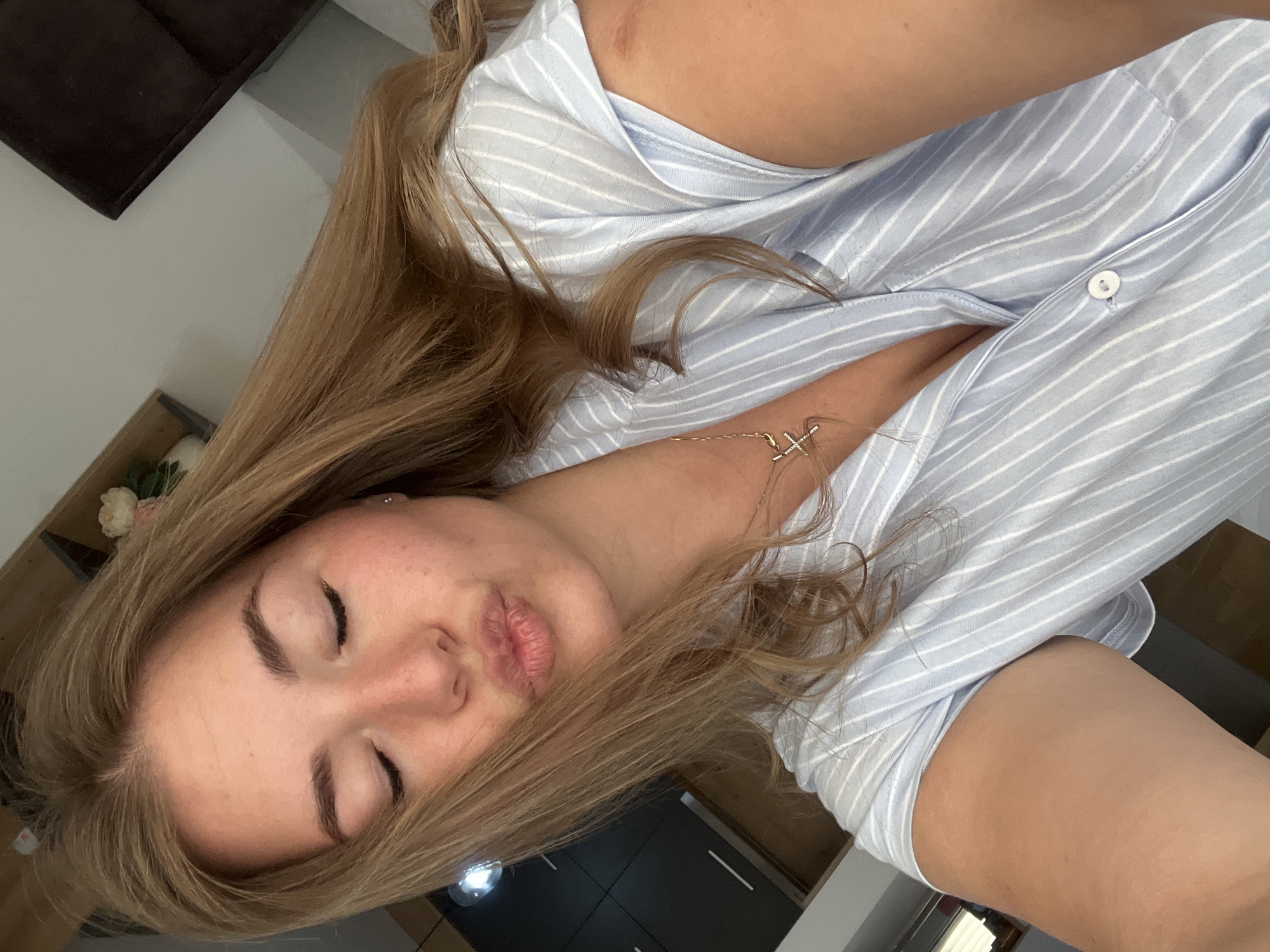 The image depicts a close-up, slightly angled indoor selfie of a female individual. She has long, light brown hair, closed eyes, and her lips are pursed in a kissing gesture. She is wearing a light blue collared shirt with thin white vertical stripes, unbuttoned to reveal a gold-colored necklace featuring an "X" shaped pendant. A faint red mark is visible on her upper right arm. The background suggests an interior space; a dark cabinet with two horizontal handles is present on the left, a light-colored wall and dark furniture are to the upper right, and a wooden shelf with floral decor is partially visible in the upper left. The specific location within the city of Budva, Montenegro, is not discernible from the visual content of the image.