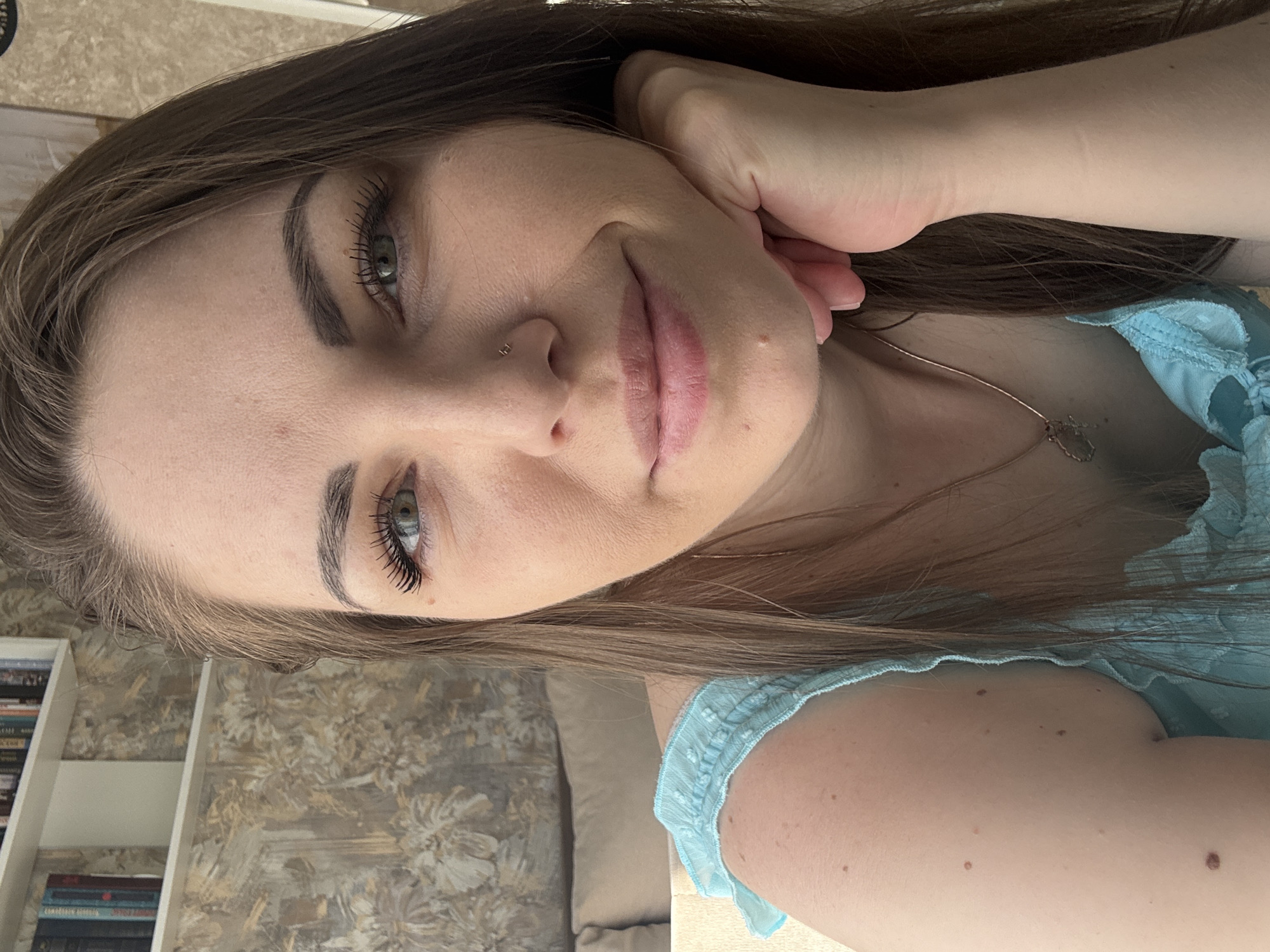 A woman with brown hair and green eyes, wearing a blue top, poses for a selfie. Her head is tilted and rests on her hand. She has a nose piercing. A bookshelf is visible in the background, and the backdrop suggests an interior setting, possibly a living room or bedroom. The location is specified as Раздолье, Russia.