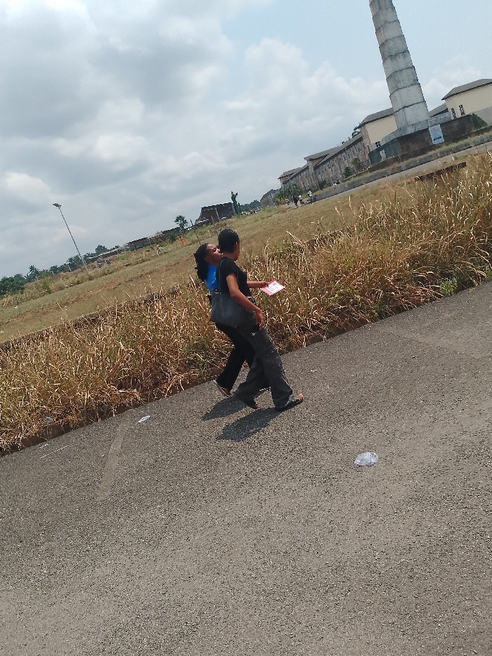 Two people walk together on an asphalt road with grassy areas on the side, near the National War Museum in Umueshi, Nigeria. The person on the left wears a blue shirt and dark pants, while the person on the right wears a black top, gray pants, and carries a dark-colored bag over the shoulder. The person on the right holds a pink object in hand. A tall chimney-like structure is visible in the background, as well as other buildings. Litter is scattered on the road.
