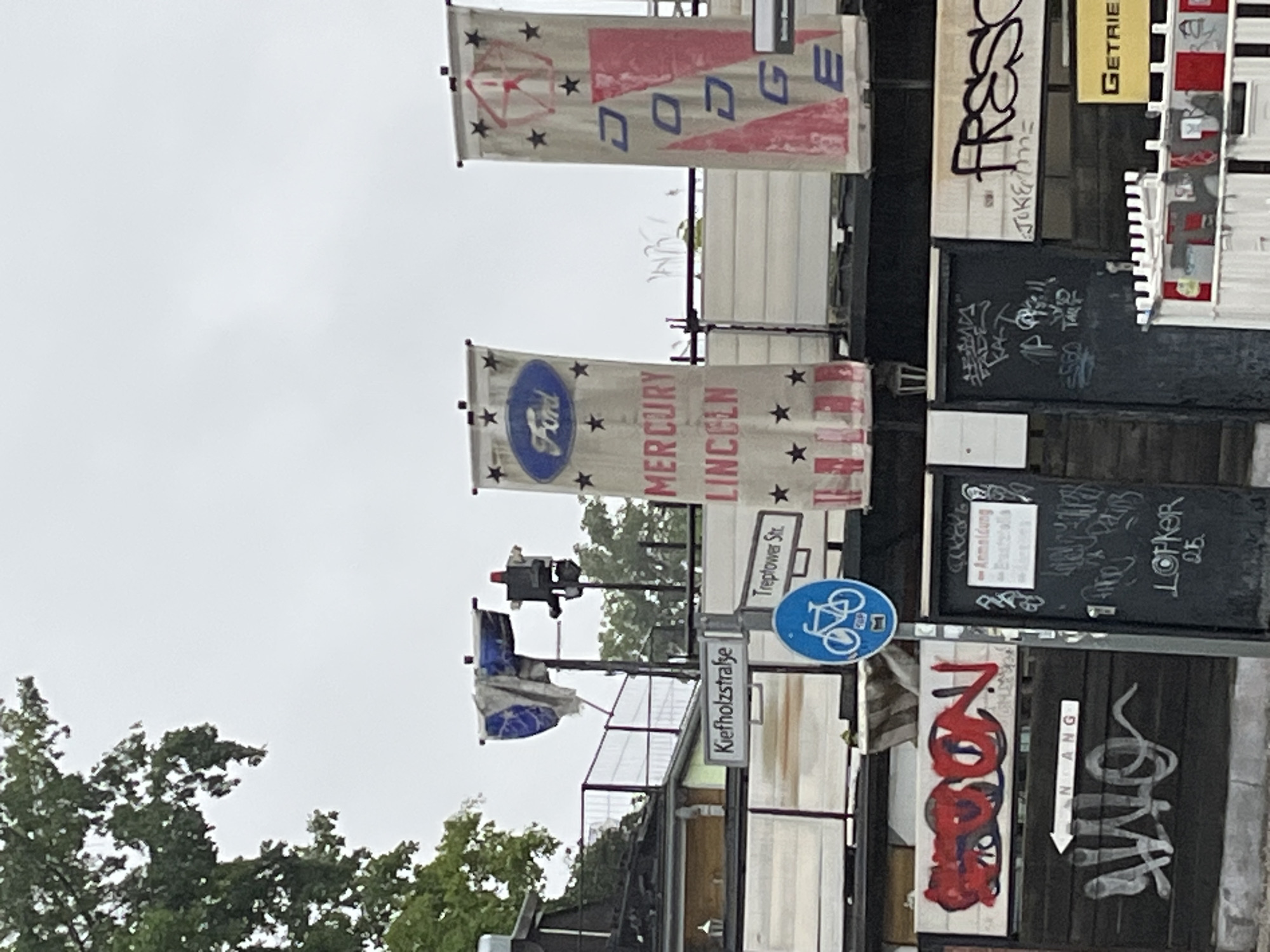 The image depicts an outdoor scene with several banners hanging, likely in a commercial or industrial area of Berlin, Germany. The banners feature logos and names associated with car brands, specifically Ford, Mercury, Lincoln, and Dodge. One banner prominently displays the Ford oval logo, while another shows the Dodge logo and name. The banners are suspended from poles, suggesting they are part of a display or advertisement.
In the foreground, there is a dark wooden fence with various graffiti tags. One tag, in red, appears to read "SPOON." Below this, an arrow points left with "N ANG" written on it. Another white graffiti tag is visible on the fence. A dark door is present to the right of the fence, also covered in graffiti, including what appears to be an "Anmeldung" (registration) sign.
A street sign indicating "Kiefholzstraße" and "Treptower Str." is visible, confirming the location in Berlin. A blue circular sign with a bicycle symbol is also present, indicating a cycling route or regulation.
Behind the fence and banners, a building or structure is partially visible, with some scaffolding and white panels. A traffic light is also seen mounted on a pole, with a red light illuminated. The overall environment appears somewhat weathered and industrial, with an overcast sky suggesting potentially cloudy weather.