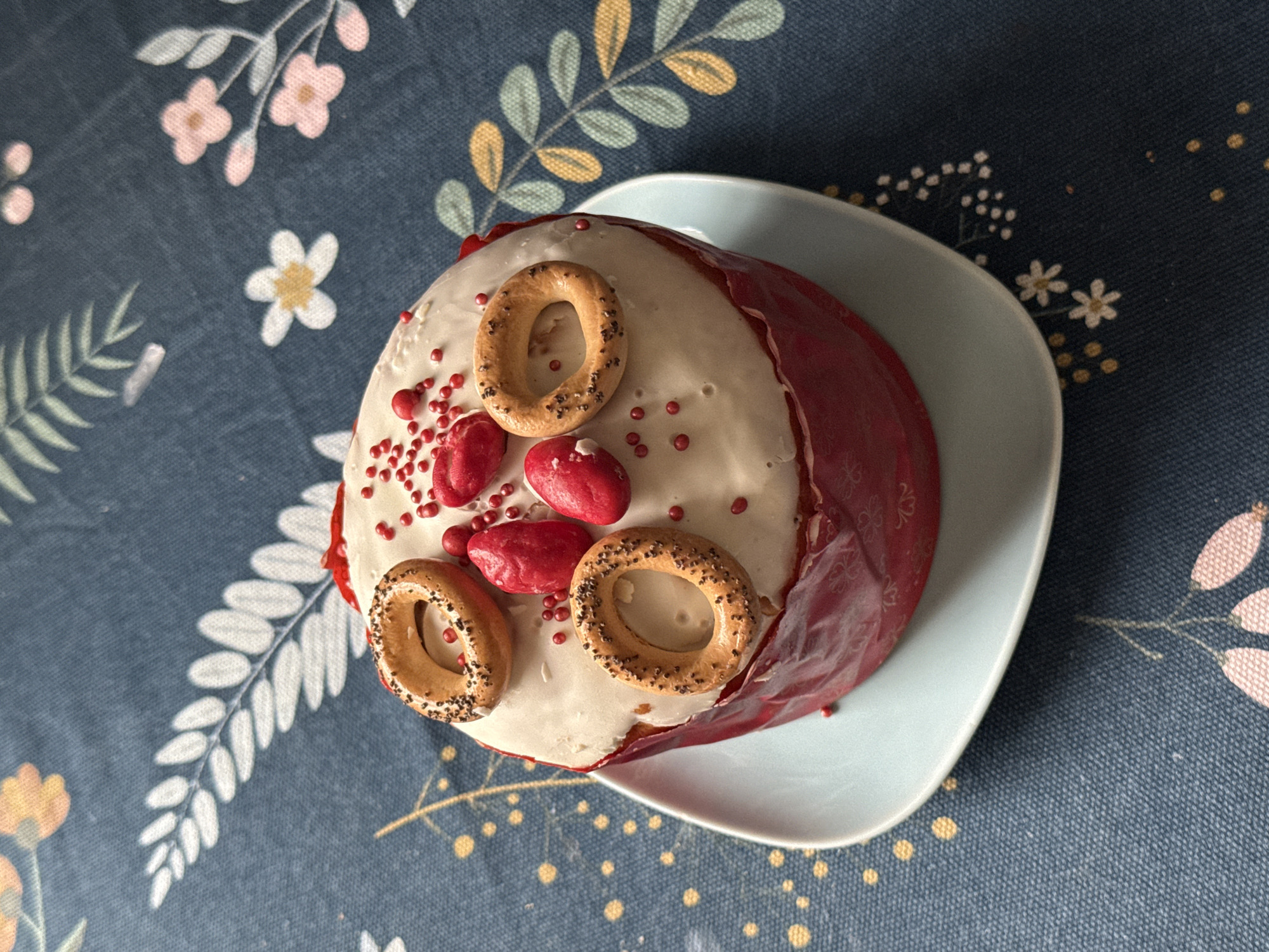 A top-down, eye-level close-up shot reveals a decorated Easter cake, known as Kulich, placed on a light blue plate. The tablecloth beneath features a floral pattern with shades of blue, pink, white, and yellow. The Kulich is adorned with white icing, small red candies, and three circular bagels. The cake's wrapper is red. The precise location within Moscow, Russia, cannot be determined from the visual information. No people or interactions are visible.