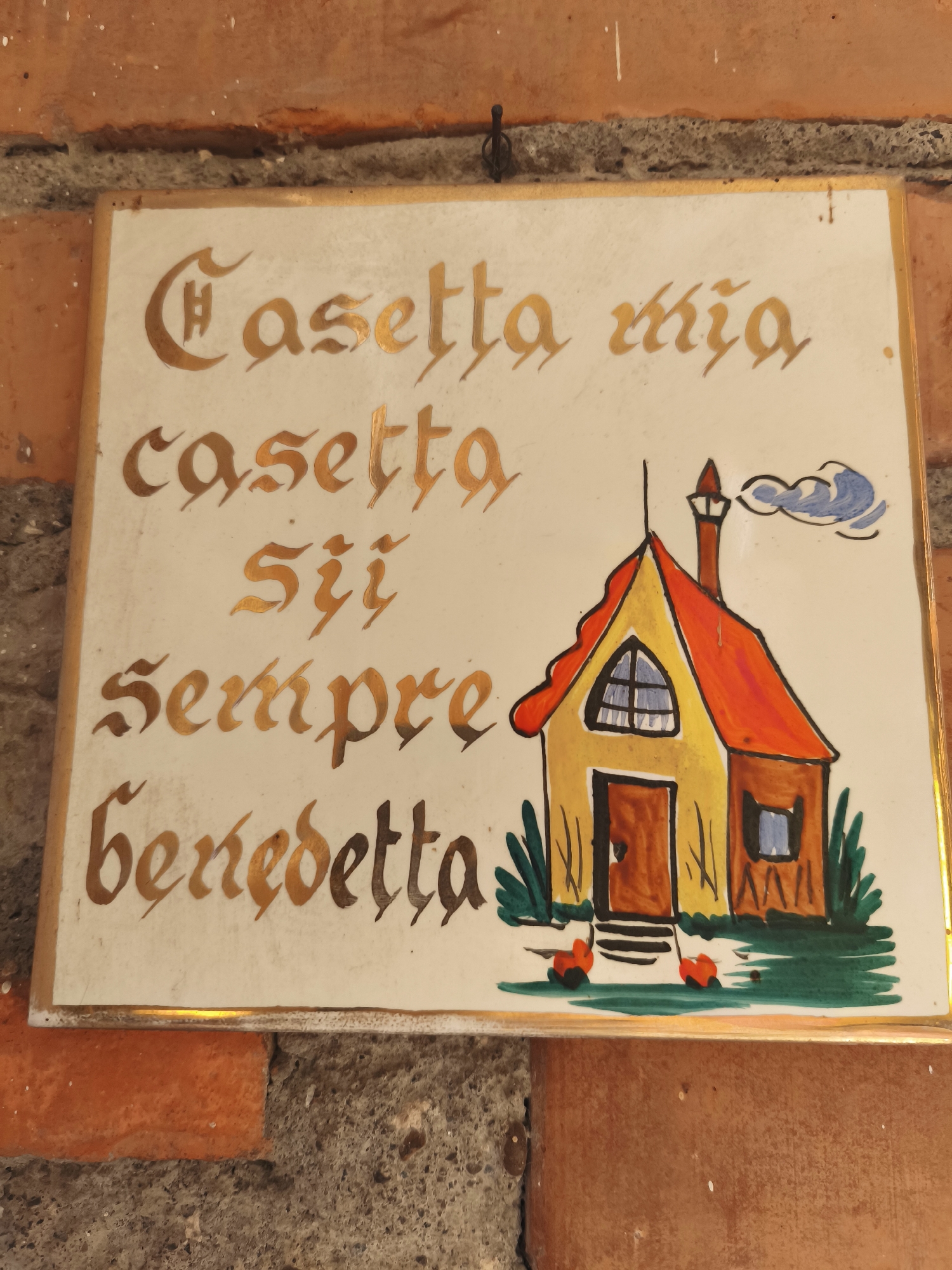 This is a close-up, eye-level shot of a decorative ceramic plaque attached to a rustic brick wall. The plaque, with a thin gold border, features a whimsical drawing of a yellow house with an orange roof, a red-orange chimney, and blue-windowed openings. Green foliage and two small red figures, possibly chickens, are depicted in front of the house. Above and to the right of the house, a stylized blue cloud drifts.
The plaque is inscribed with golden calligraphy. The text reads: "Casetta mia casetta sii sempre benedetta". This translates from Italian to "My little house, little house, may you always be blessed." The style of the lettering is ornate and flowing, adding to the charming, old-world feel of the plaque. The setting appears to be outdoors, given the visible brickwork, suggesting it might be mounted on an exterior wall of a home or building. The lighting suggests it is daytime, with no specific indicators of time of day or weather. The overall impression is one of warmth, domesticity, and good wishes. The location context provided is Passignano sul Trasimeno, Italy, which aligns with the Italian text on the plaque.
