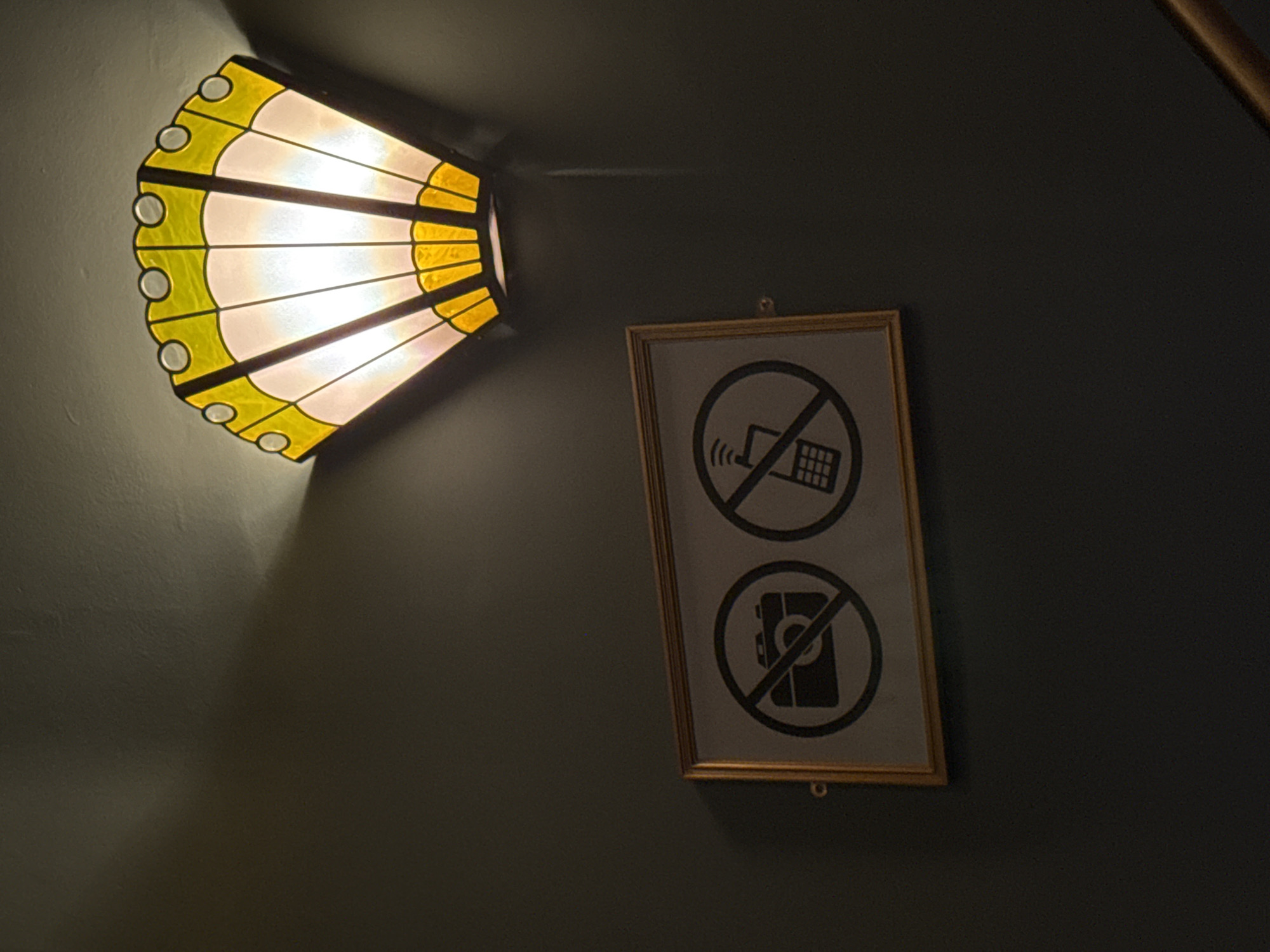 A dimly lit interior scene featuring a wall with a decorative, semi-circular, stained-glass-style lamp on the left side and a framed sign on the right. The lamp emits a warm glow. The sign contains two symbols: an image of an older style cell phone (likely a flip phone) with radio waves emanating from it, and a second symbol displaying an image of a digital camera; both of these images are crossed out with a prohibition symbol (a circle with a diagonal line). The sign likely indicates that cell phone use and photography are prohibited in this location.
While it is impossible to confirm with the existing image, the framed sign may indicate this image was taken in a theater or cinema in London.