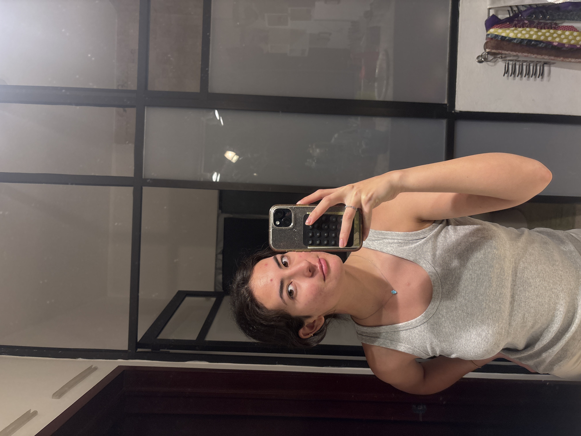 A woman is taking a selfie in a room in Puebla, Mexico. The woman has dark hair and is wearing a grey tank top and a gold necklace with a blue stone. She is holding a gold-colored phone in her right hand. The room has a large window with black frames. Behind the window, there are other buildings and possibly a bicycle. To the right of the window is a white wall with a clothing rack. The floor is made of dark wood.