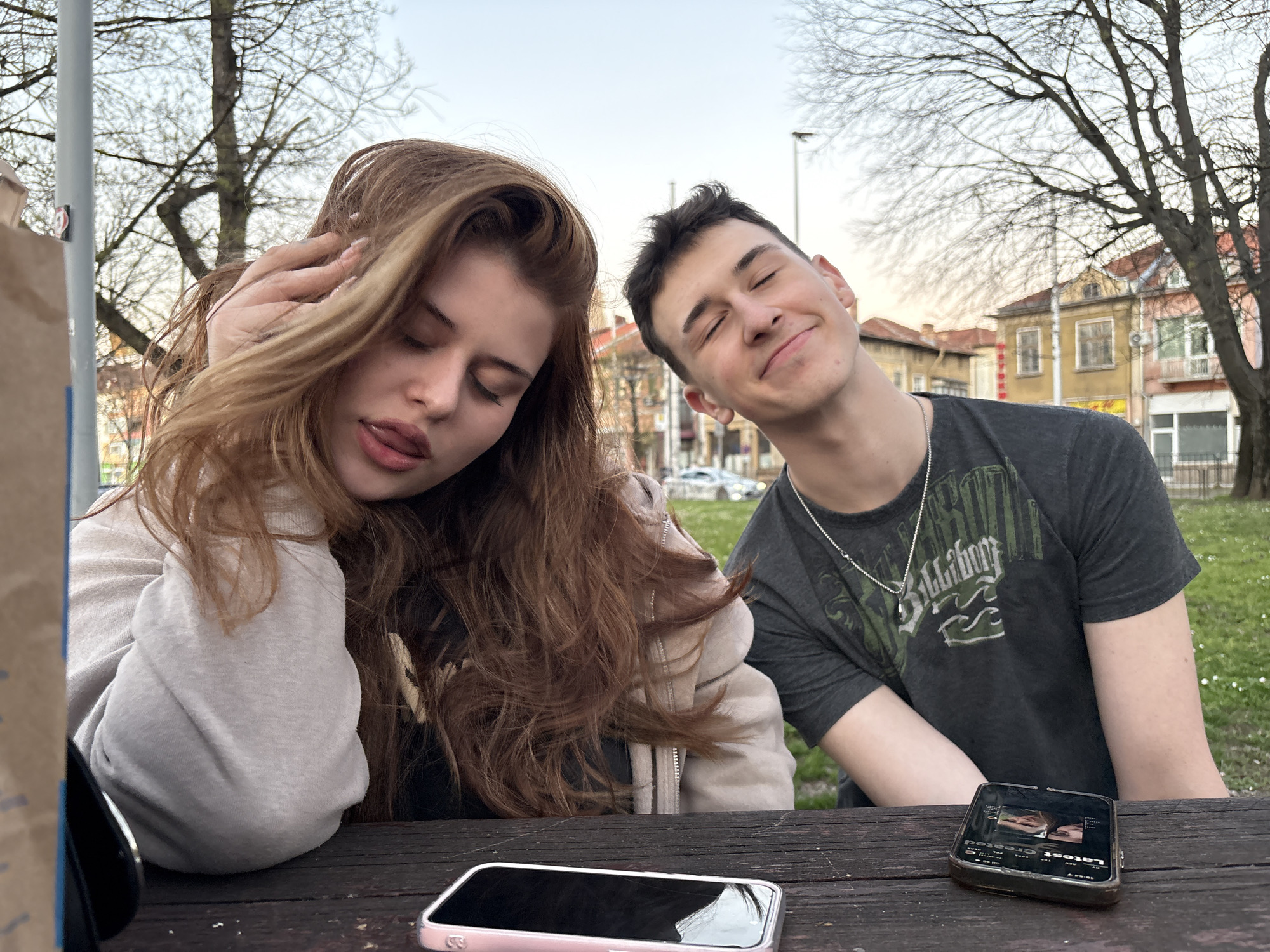 The image depicts two young adults sitting at a wooden table outdoors. The woman on the left has long, wavy brown hair and is wearing a light-colored hooded jacket. Her eyes are closed, and her tongue is slightly visible. The man on the right has short, dark hair, closed eyes, and a slight smile. He's wearing a dark grey t-shirt with a Billabong logo and a silver necklace. Two smartphones are lying on the table, a pink one and a black one, as well as a small outdated portable TV/Radio on the corner of the table.
The background shows a park-like setting with bare trees and buildings. The buildings are identifiable as being located in Pleven, Bulgaria due to the distinct architectural style of the buildings. It is daytime, and the sky is slightly overcast.