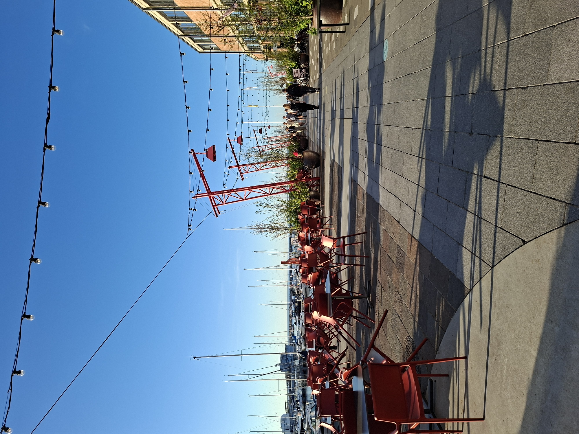 This image depicts a vibrant waterfront promenade located in the Noblessner Quarter of Tallinn, Estonia. The scene is illuminated by clear sunlight, casting long shadows from left to right, suggesting a low sun angle, either morning or late afternoon, under a clear blue sky.
On the far left, a marina is filled with numerous sailboats and yachts, their white masts sharply silhouetted against the blue sky. The water in the marina appears calm.
Immediately to the right of the marina, a large outdoor dining area occupies the left half of the promenade. It features multiple dark tables and numerous bright red chairs. Many chairs are stacked on tables or overturned, indicating the area is either preparing for business, closed, or not currently occupied by diners. The ground in this section is paved with reddish-brown cobblestones.
Running parallel to the dining area, a wide pedestrian walkway, paved with light grey rectangular tiles, extends into the distance on the right. Several individuals are walking along this path. In the mid-ground, two people, dressed in dark clothing, walk away from the viewer. Further down the promenade, additional people are visible.
Architecturally distinct, red, angular metal structures, serving as supports for overhead lighting, are interspersed along the walkway. Strings of small, clear light bulbs are suspended between these red structures and the adjacent building, as well as strung across the sky over the dining area. Large, dark planters containing small trees or shrubs are placed along the promenade.
On the right side of the image, a multi-story building constructed of orange-brown brick with numerous windows forms the backdrop to the promenade. Its industrial-chic design is characteristic of the converted shipyard buildings in the Noblessner area. A sign, partially visible, appears to read "Kunst ring".