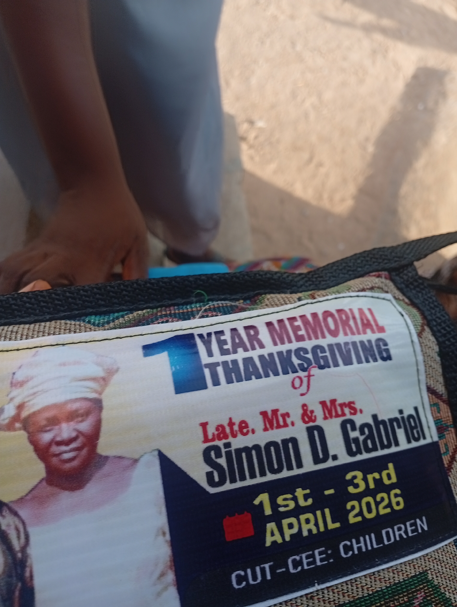 This image shows a close-up of a memorial bag with a portrait of a woman wearing a headscarf and a white garment. Text on the bag reads "1 YEAR MEMORIAL THANKSGIVING of Late. Mr. & Mrs. Simon D. Gabriel 1st - 3rd APRIL 2026 CUT-CEE: CHILDREN". The bag has a woven fabric pattern and a black handle. The background is out of focus and appears to be an outdoor setting with sandy ground and shadows, possibly indicative of a public space or gathering. The image does not provide information to determine a specific location within Jalingo, Nigeria.