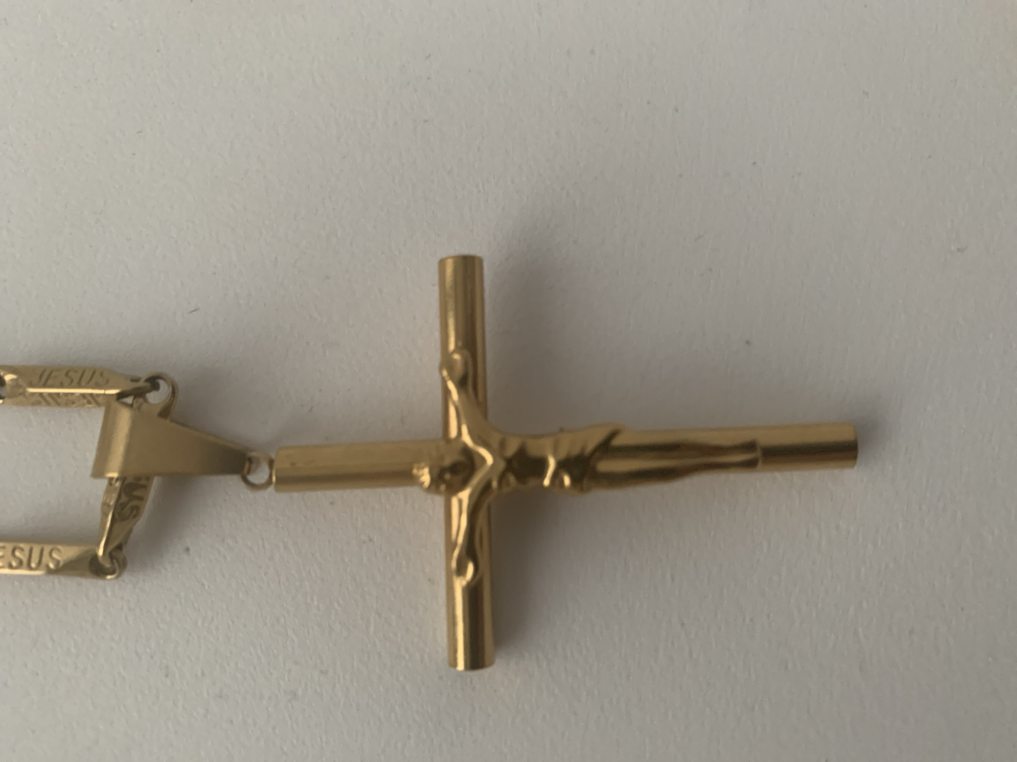 A gold crucifix pendant hangs from a gold chain. The crucifix depicts Jesus in a stylized, elongated form. The chain appears to be made of interlocking rectangular links and has the word "JESUS" inscribed on some of the links. The background is a plain, lightly textured white surface, likely a wall or table. The lighting is even, casting soft shadows. The image is a close-up shot, focusing on the pendant and a small portion of the chain. There are no people or other objects visible in the frame.
