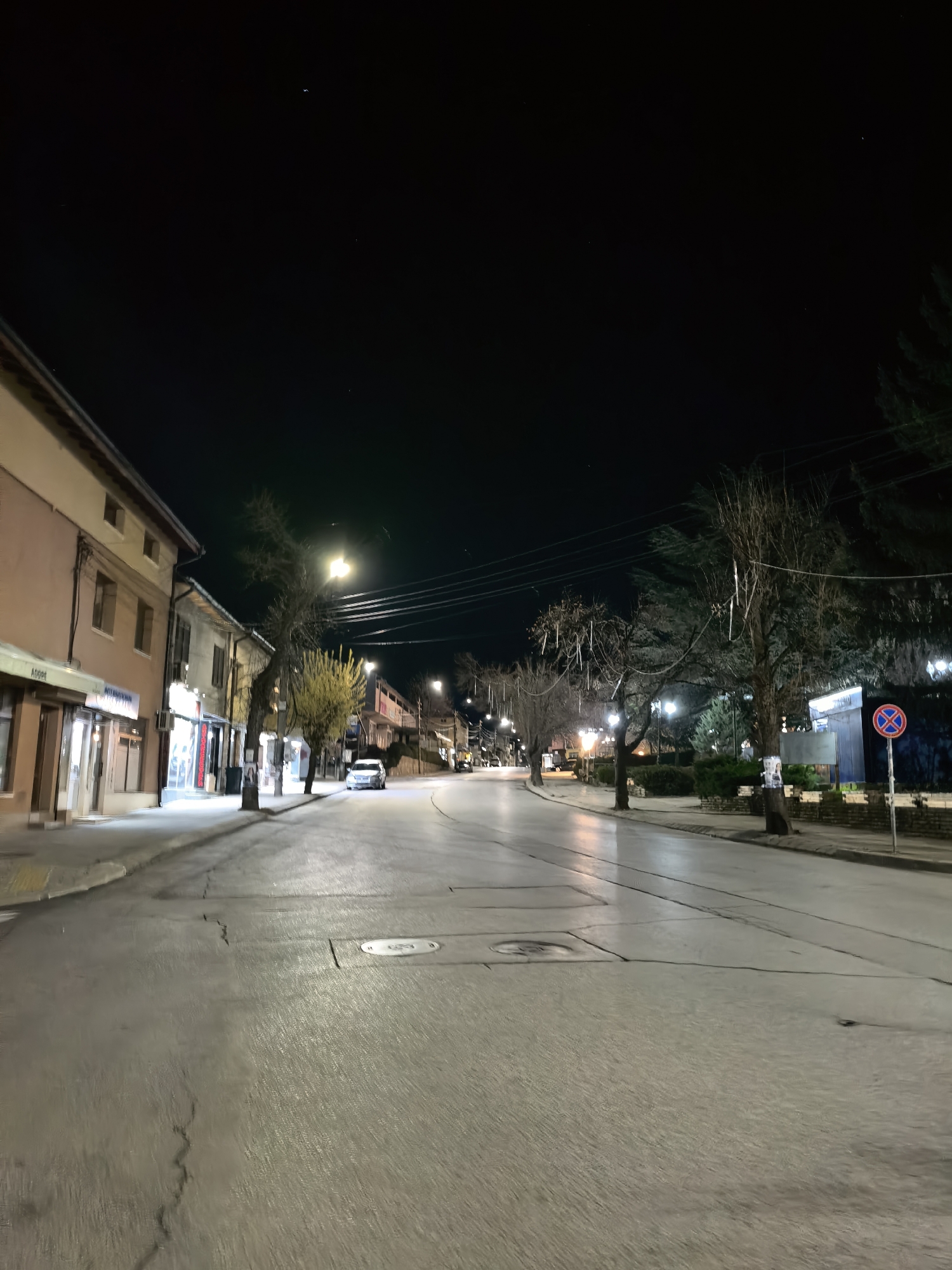 The image depicts an empty street at night in a city. The location is characterized by two-story buildings on the left side, illuminated by streetlights and storefront lights. There are bare trees and evergreen trees planted along the right side of the street, with a no-stopping sign visible. The street is paved and marked with lane dividers, exhibiting some cracks and manhole covers. In the distance, there are more buildings and streetlights fading into the night. The sky is dark, with a few visible stars. No people are visible in the image.