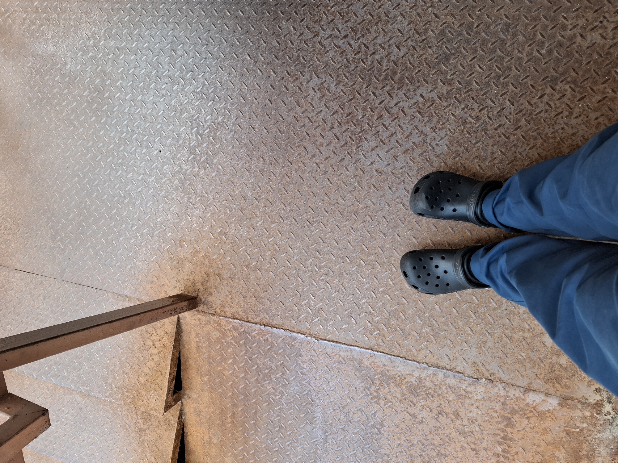 The image shows a pair of legs wearing blue pants and black Crocs, standing on a diamond plate metal surface. The surface appears to be part of a stairway, with a metal railing visible on the left side. The metal has a worn and slightly rusty appearance. The perspective is from above, looking down at the person's feet and the stairs. The lighting suggests it could be daytime, but there are no clear indicators of the weather or a specific time of day. The scene is simple and focuses on the footwear and the textured metal surface. There is no visible text in the image. The location context of Grigorovo, Russia, is provided but not visually evident from the image itself.