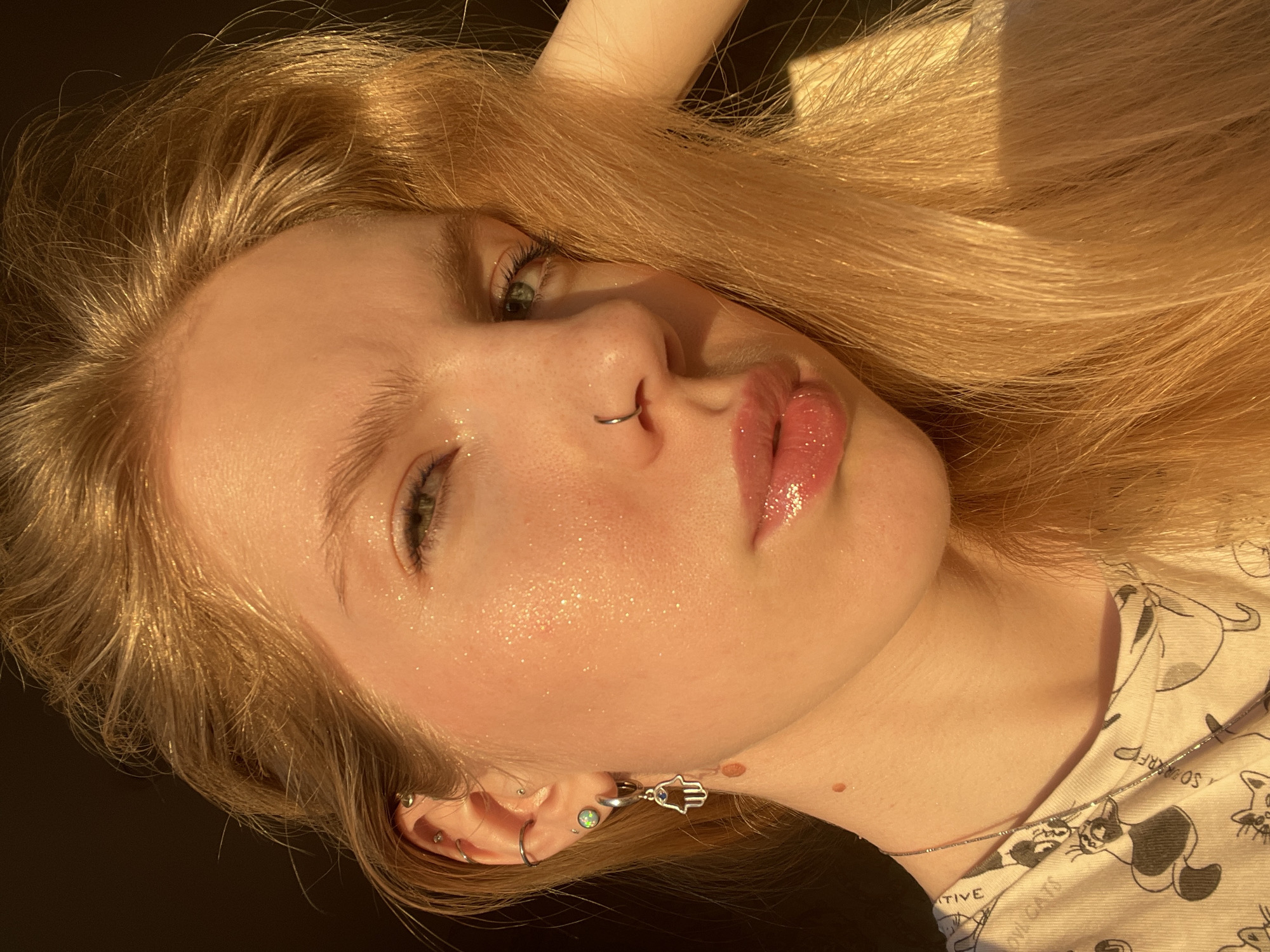 A close-up portrait depicts a young woman with long, blonde hair and fair skin. Her face is angled, revealing her facial features in detail. She has green eyes, a nose ring, and glossy lips. Light is hitting her face, creating a shimmering effect on her skin. Multiple piercings are visible in her left ear. She's wearing a t-shirt patterned with cartoon cats and a thin necklace. The background appears dark and out of focus. There is no evidence to suggest the image was taken in Izhevsk, Russia.