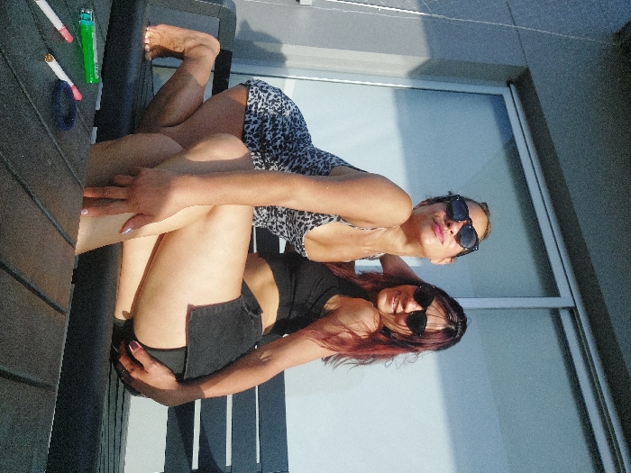 Two women pose in front of a window. One woman wears a spotted top and sunglasses. The other woman wears a dark tank top, a dark skirt, and sunglasses, with reddish-brown hair. They are on a patio or balcony. A pack of cigarettes, a lighter, and a hair tie are on a table. The sun is shining brightly.