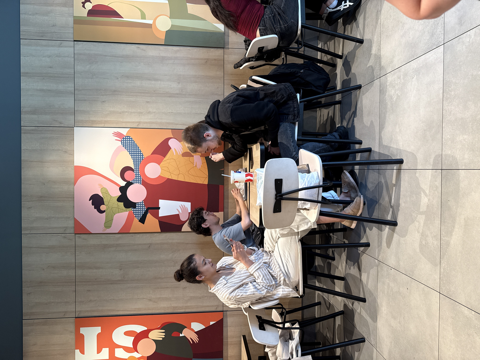 Four individuals are seated at tables in a brightly lit indoor establishment, likely a fast-food restaurant, based on the artwork and table settings. The environment features light wood-paneled walls with vibrant, abstract murals depicting stylized figures. The floor is tiled in a neutral gray.
On the left, a woman with her hair in a bun is seated, wearing a striped button-down shirt and light-colored pants. She is looking down at her mobile phone, which she holds in her hands. In front of her is a white cup with a red lid and straw.
To her right, a man with dark curly hair and glasses is seated. He is wearing a gray t-shirt and is looking down at something on the table in front of him.
Further to the right, a man with short blonde hair is seated, wearing a dark hooded jacket over a dark t-shirt and jeans. He is looking to his left, with one hand near his mouth. In front of him is a white cup with a red lid and straw. A black backpack is visible on the floor behind him.
On the far right, partially visible, is an individual with long dark hair, wearing a red top and dark jeans. They are seated and appear to be looking down.
The tables are simple, white-topped surfaces, and the chairs are modern with white seats and black legs. The overall scene suggests a casual dining environment.
The provided location is Izhevsk, Russia.