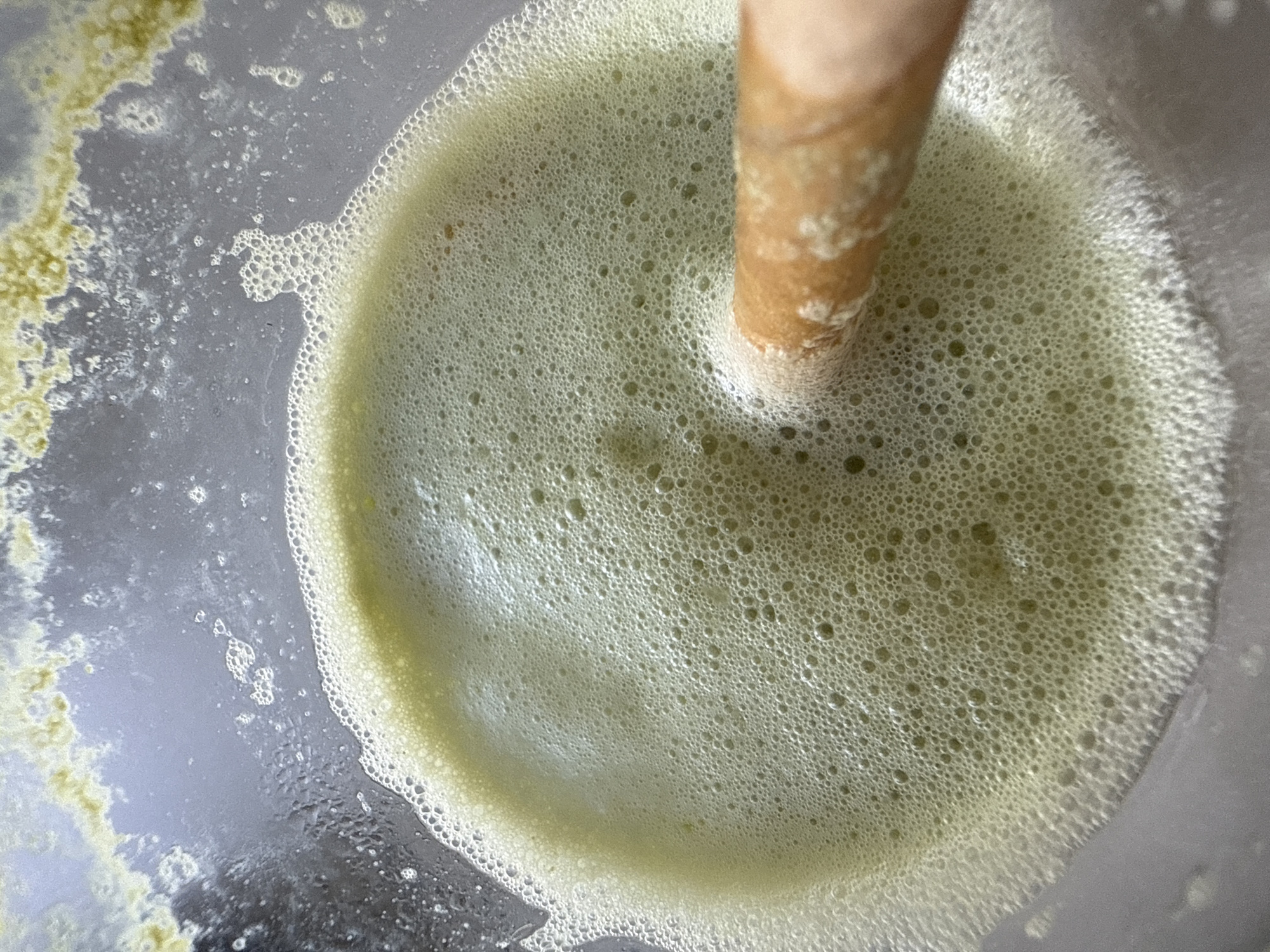 A close-up, overhead shot shows a pale green, foamy liquid in a clear container, likely a blender or a glass. The liquid has a dense layer of small bubbles across its surface, with some larger bubbles near the edges. A light brown, cylindrical object, possibly a tamper or a spoon, is partially submerged in the liquid, creating ripples and disturbing the foam. The background is blurred and appears to be the interior of the container, with some yellow-green residue clinging to the sides. There is no discernible indication of a specific location in North Kuta, Indonesia. The image focuses solely on the texture and appearance of the foamy liquid and the object within it.
