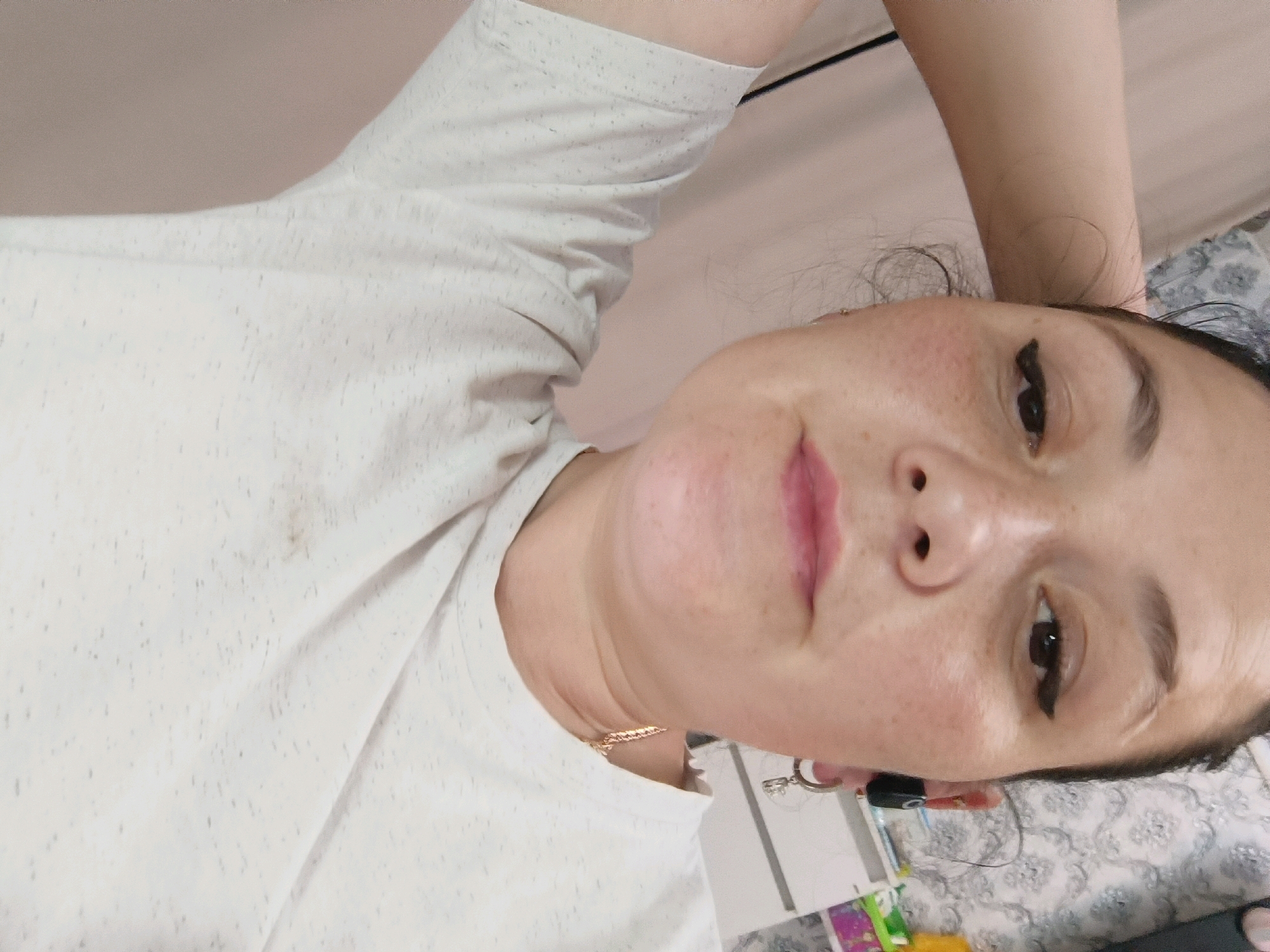 A close-up, overhead, slightly blurry, selfie shows a person lying down with their arm raised behind their head. The person is wearing a white t-shirt with a speckled pattern. Their face is partially visible, with their eyes open and looking towards the camera. They have makeup on, including eyeliner and lip gloss. The background is out of focus and appears to be indoors, possibly a bedroom, with a light-colored wall and a patterned blanket or sheet. There are some small objects visible on a surface to the right of the person, including what appear to be a pen and a keyring. The image is of poor quality and does not provide enough detail to determine the location within Chaikovsky, Russia, or any specific actions or interactions.