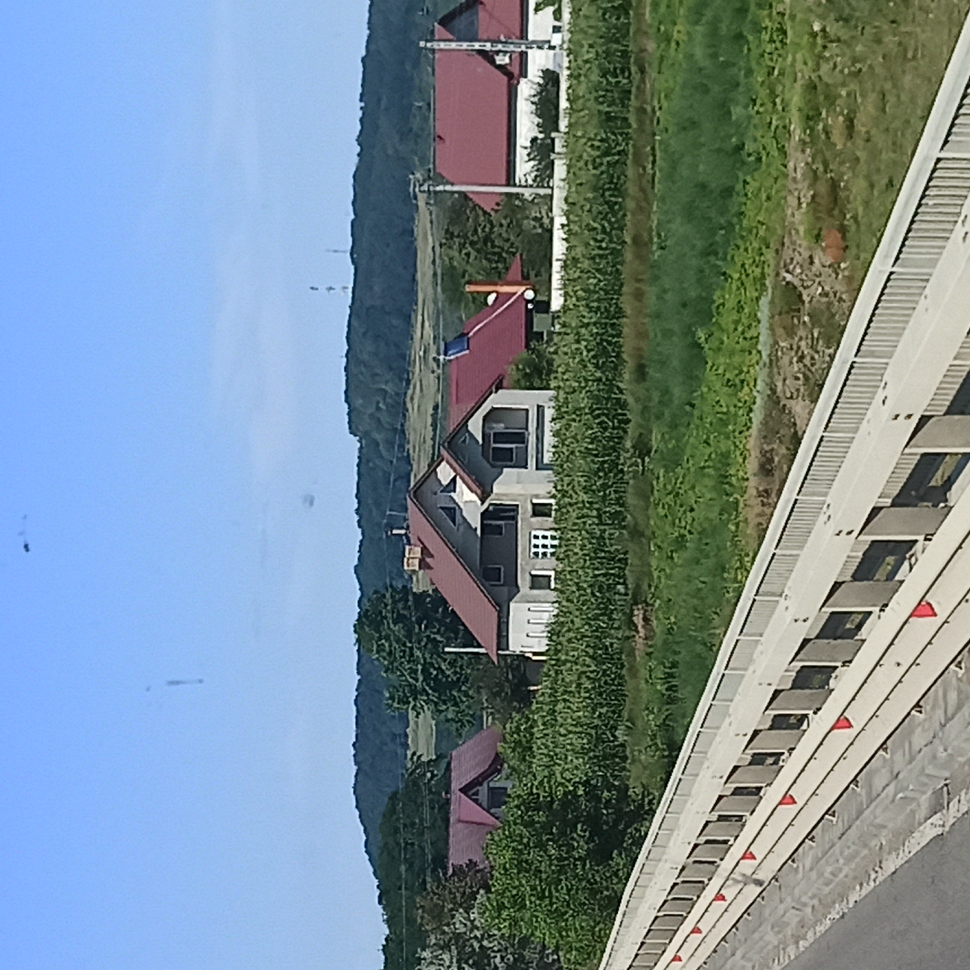 The image depicts a rural scene with several houses in the background, situated amidst greenery and a cornfield. In the foreground, there is a bridge or overpass with a white railing. The sky is clear and blue. The houses appear to be modern, with red tiled roofs. One house is particularly prominent, featuring a complex roofline with multiple gables and triangular windows. To the right, another house with a red roof and white walls is visible. The surrounding landscape is lush, with trees and crops. In the distance, hills can be seen. The overall impression is one of a tranquil countryside setting.