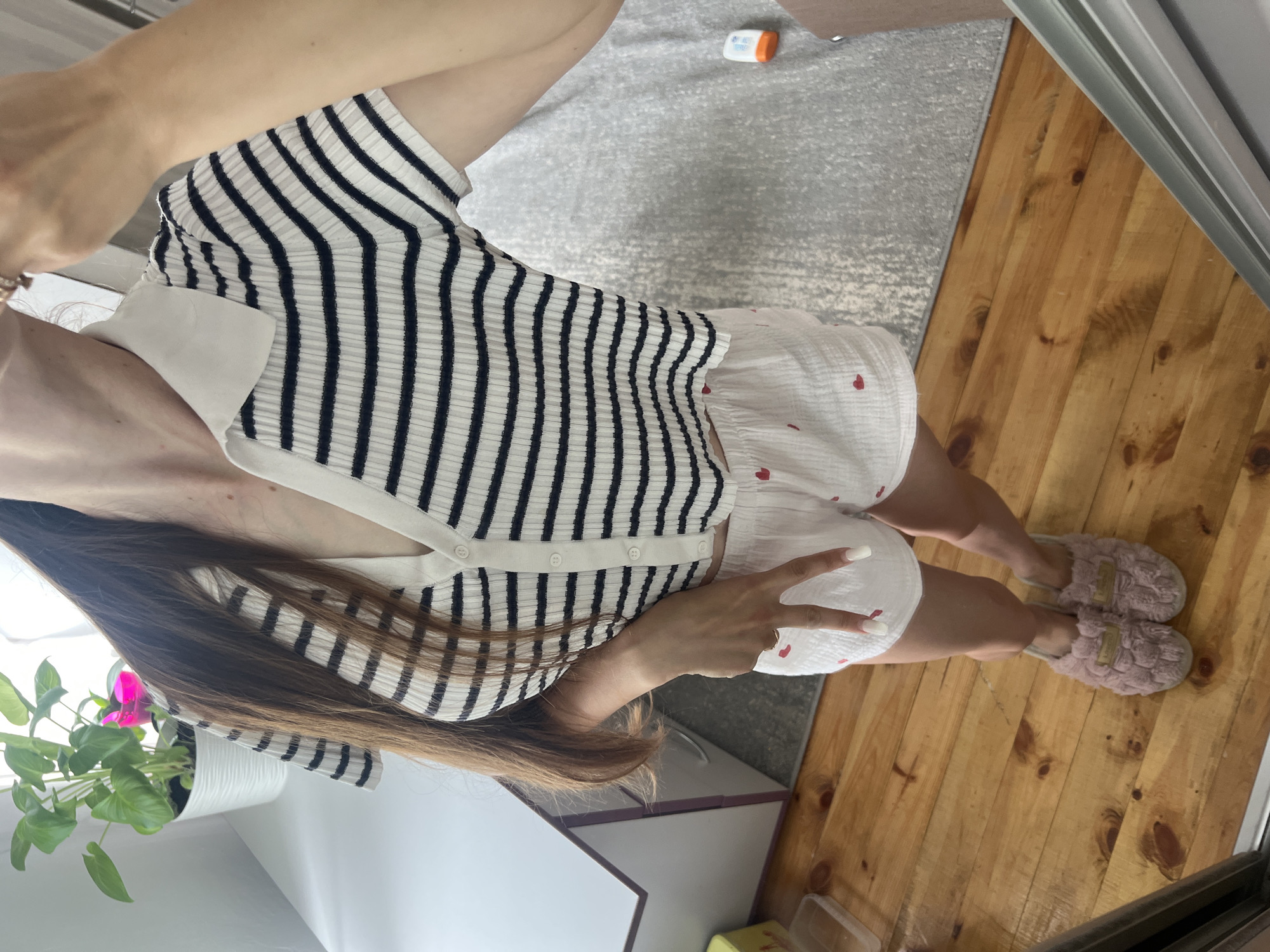 A woman stands indoors, reflected in a mirror. She is wearing a striped short-sleeved collared shirt with white and black stripes, and white shorts with small red heart patterns. She has long brown hair and is wearing pink fuzzy slippers. Her left hand is raised, making a peace sign. The background includes a potted plant on a white surface, a wooden floor, and a grey rug. The environment appears to be a room in a residence.