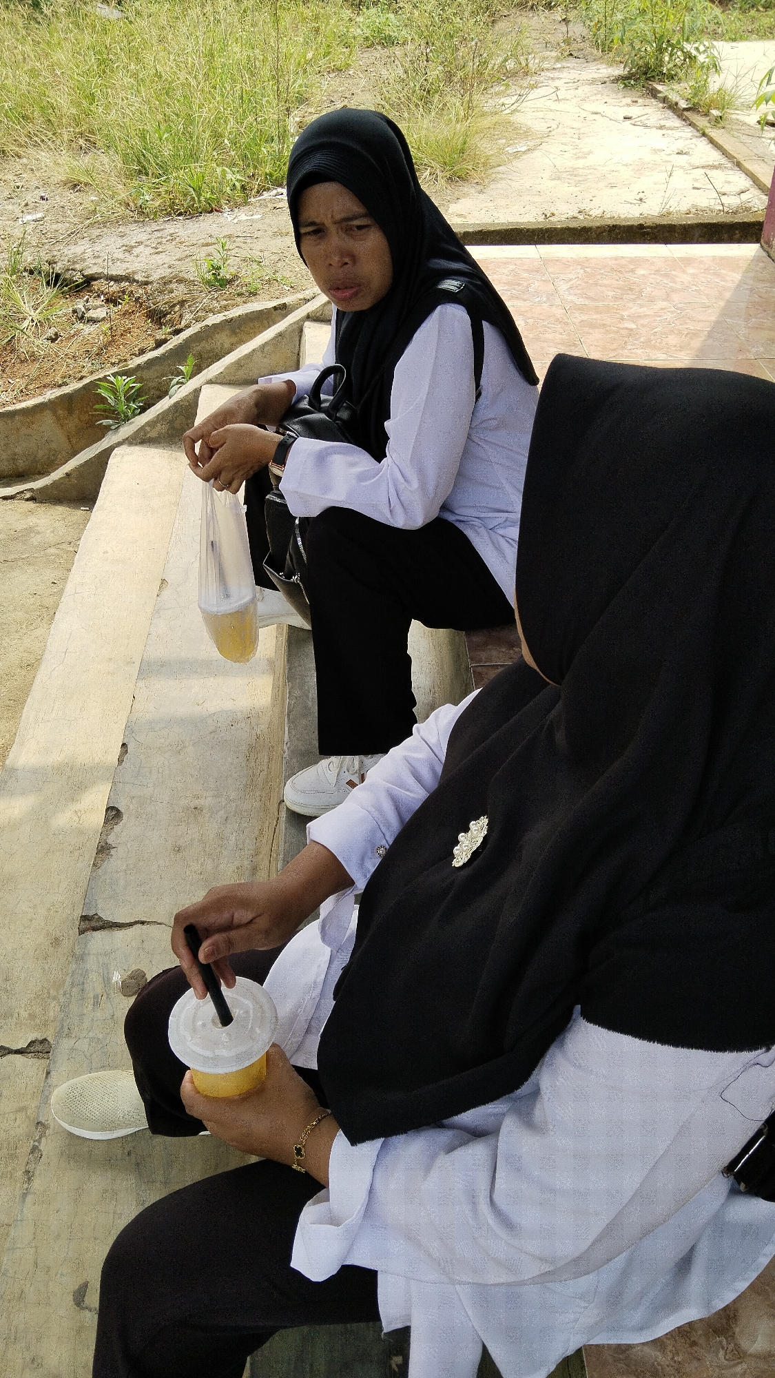 Two women in black hijabs and white long-sleeved tops are sitting on concrete steps outdoors, possibly in Margatunggal, Indonesia. The woman on the left, facing the viewer, is holding a clear plastic bag filled with a yellow beverage and is looking away with a concerned expression. The woman on the right, facing away from the viewer, is holding a yellow cup with a straw and is wearing a decorative brooch on her hijab. The background consists of dry grass, dirt, and a paved area.